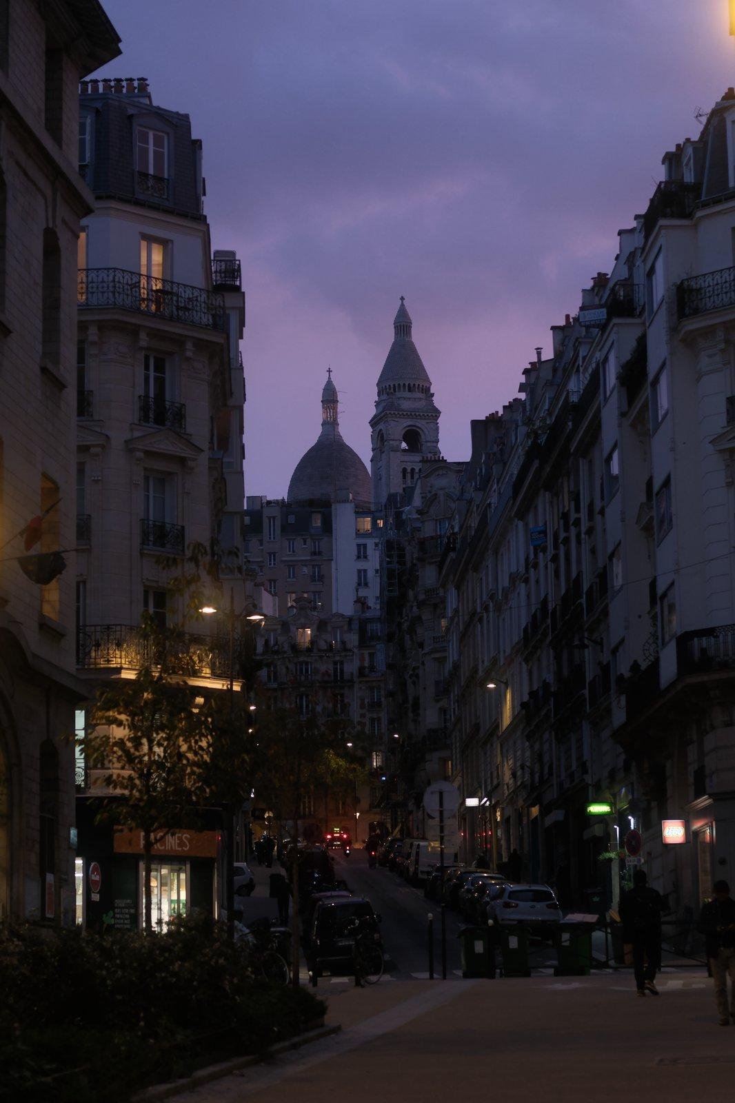 Photo verticale. Au premier plan, une fin de zone piétonne, montant vers la rue
encadrée d’immeubles parisiens qui sont surplombés d’un ciel violet / bleu, avec
le haut du Sacré-Cœur qui pointe