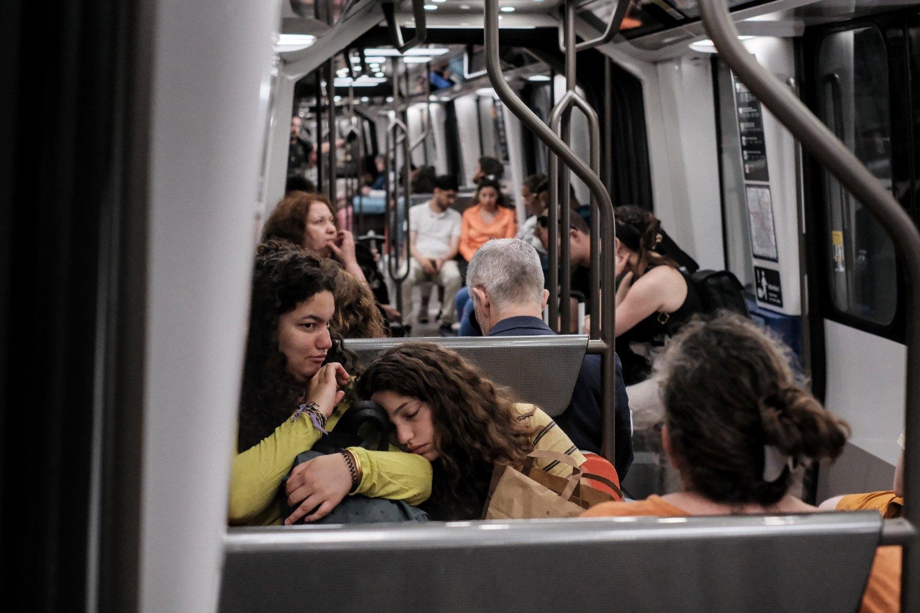 Photo horizontale. Dans un wagon de métro, assises dans un carré de 4, une jeune fille au cheveux bruns ondulés repose sa tête sur l’épaule de sa sœur