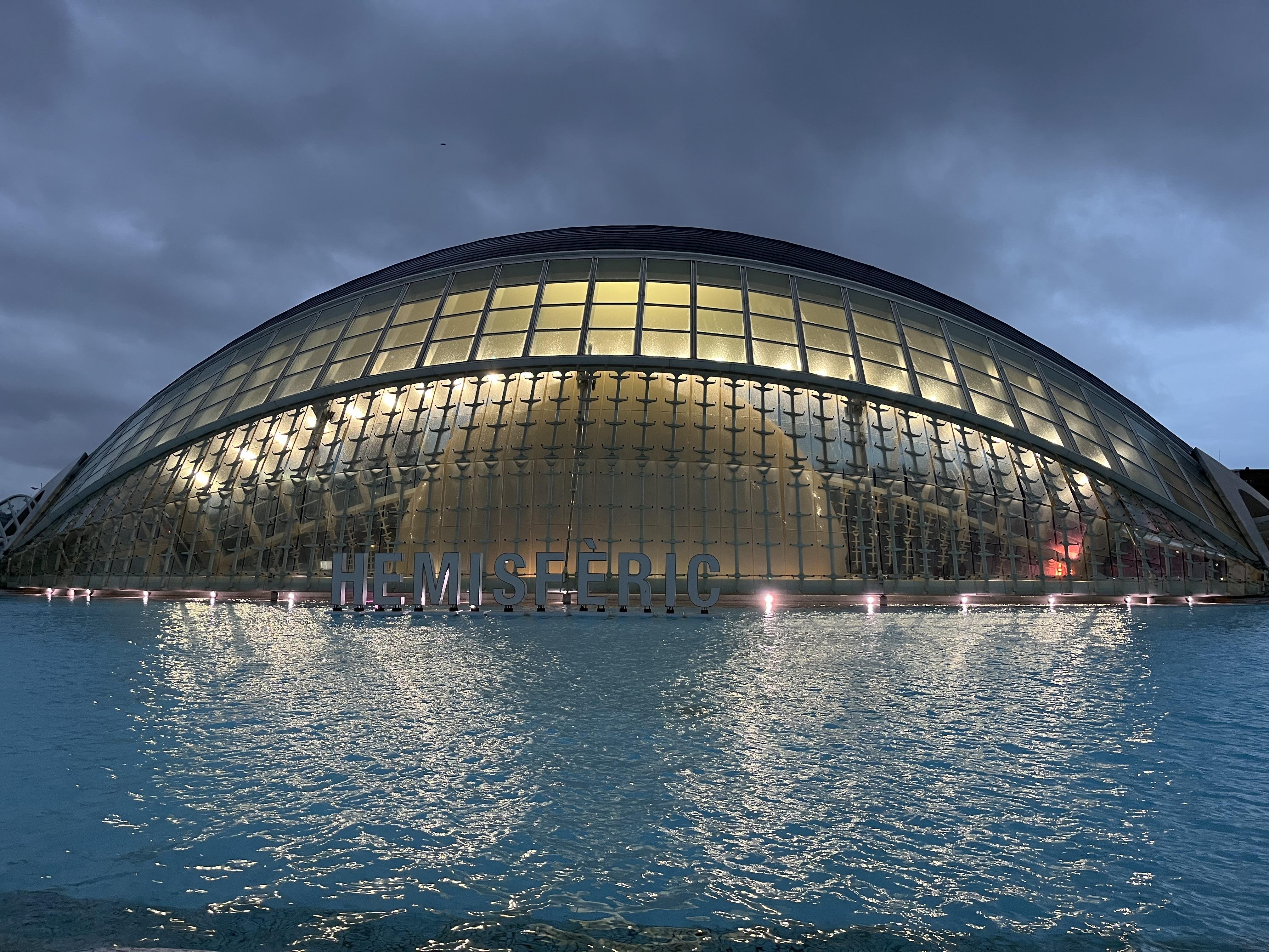 The image shows the L'Hemisfèric, a striking architectural structure located in the City of Arts and Sciences in Valencia, Spain. It is designed to resemble a giant eye and is surrounded by a pool of water, which enhances its appearance. The building houses an IMAX cinema, planetarium, and laserium. The lights are on, creating a dramatic effect against the cloudy sky.