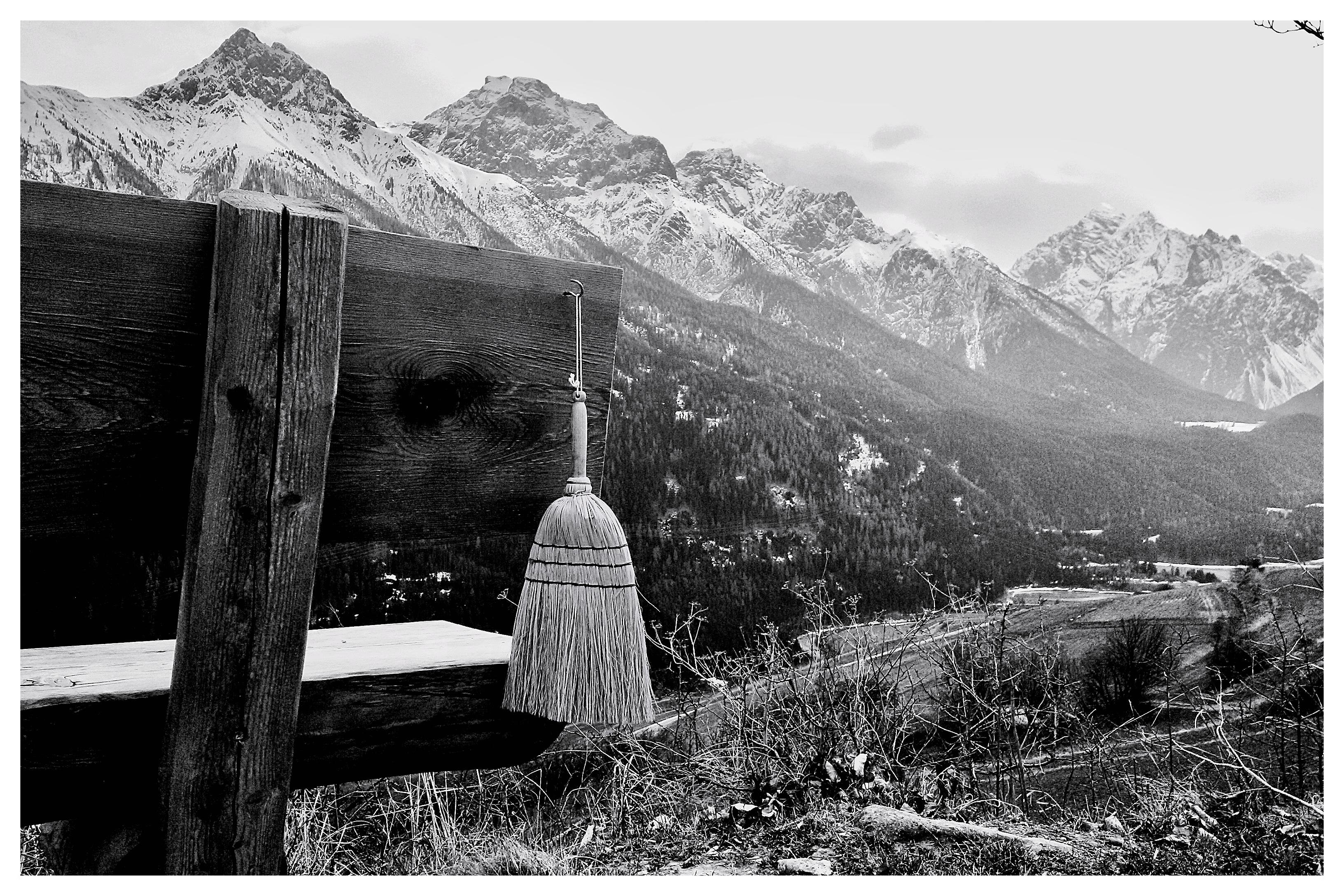 Wooden bench positioned on a hillside overlooking a vast valley with snow-capped mountains in the background. A broom is hanging from the bench. The foreground includes sparse vegetation, and the scene is in black and white, emphasizing texture and contrast.