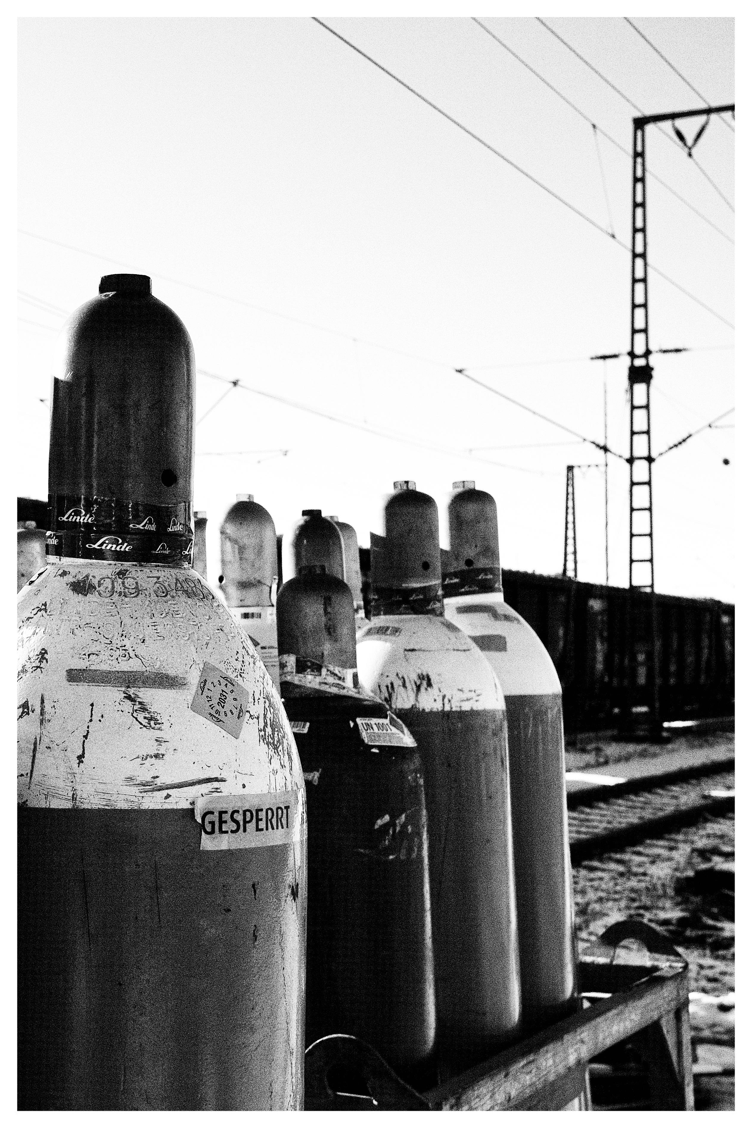 Black and white image of several large gas cylinders standing upright on a platform. The cylinders have visible labels and stickers; one prominently displays the word "GESPERRT." Overhead, railway power lines stretch across the sky, with a railway structure in the background. The ground appears textured, possibly gravel or industrial terrain.