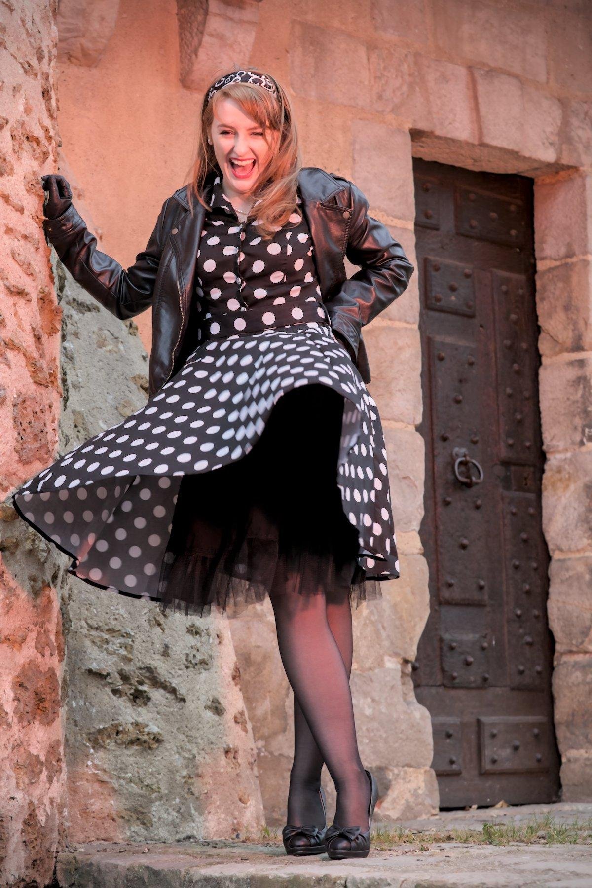 Photo verticale. Une jeune femme rousse aux cheveux long, coiffée d’un bandeau à la mode année 50, noir et blanc, une robe noire à gros pois blanc, des chaussures noires à talon haut, un blouson noir cuir et des gants noirs cuir. Le vent souffle sa robe révélant les voiles noirs dessous et lui soutirant un grand éclat de rire