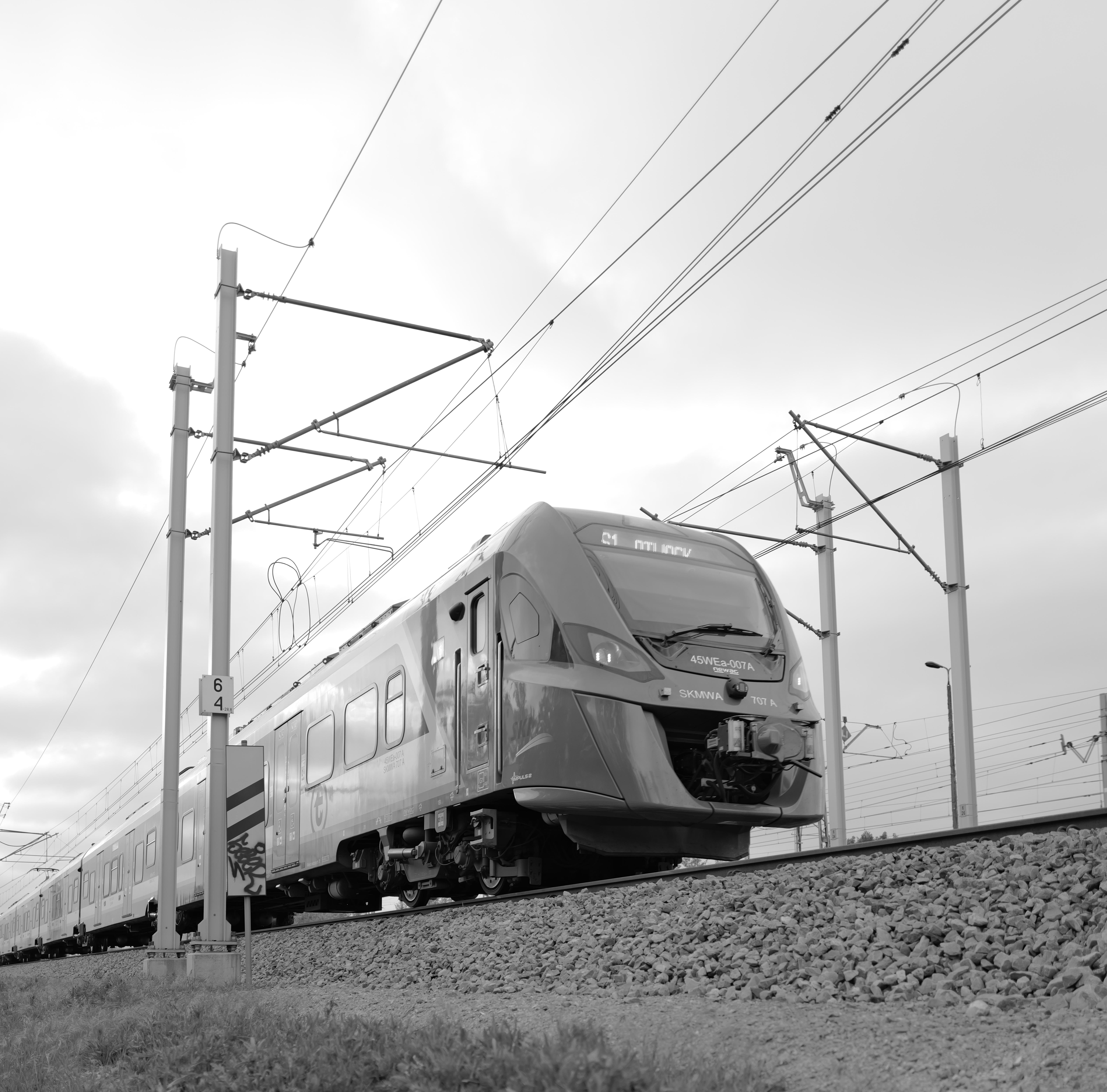 Low-angle black-and-white view of a modern passenger train on an elevated track, with overhead power lines and support poles stretching into the cloudy sky.