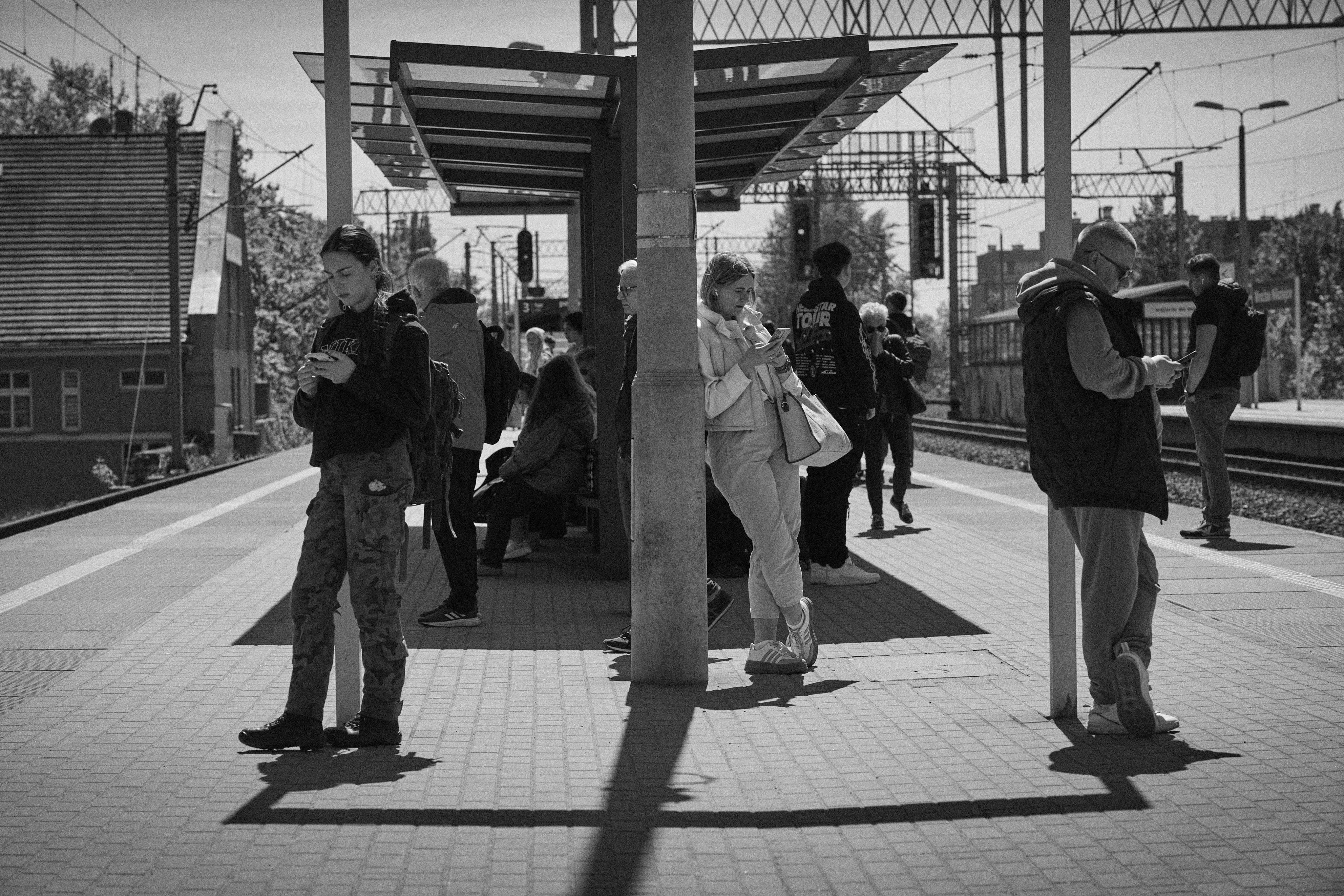 Black-and-white photo of a train station platform with a covered shelter at center.Several people stand or sit beneath it, most looking at smartphones. Train tracks run along the right side, with overhead power lines and signal lights in the background; long shadows stretch across the tiled platform.