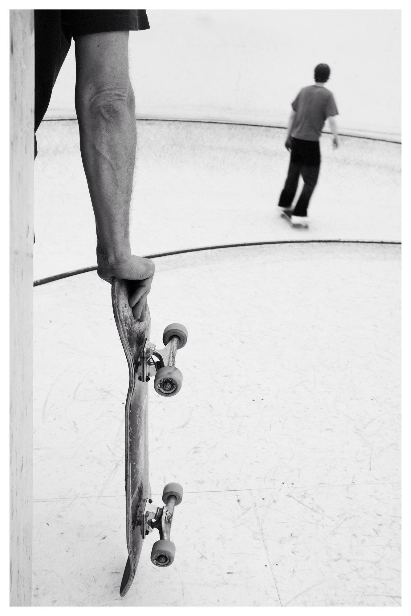 Black-and-white photo of a forearm holding a skateboard vertically in the left foreground, with a blurred skateboarder riding inside a smooth concrete skate bowl in the background.
