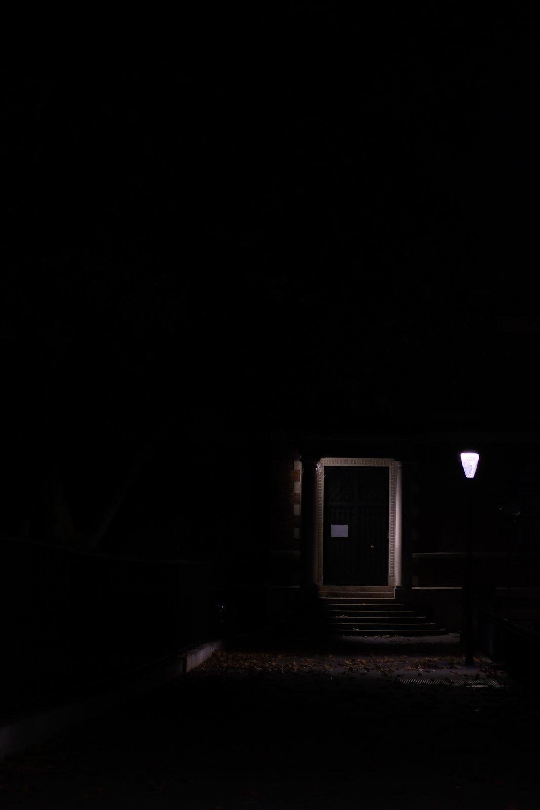 Photo format portrait, prise de nuit, seul le quart bas droite montre quelque chose. Une porte noire, faiblement éclairée par un petit lampadaire à droite de l’image. Une volée de marches en arc de cercles mène à la porte, le sol est couvert de feuilles mortes.