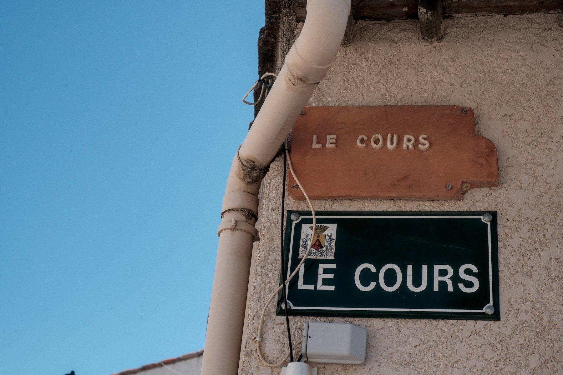Photo horizontale. Au coin d’une maison, 2 plaque donnant le nom de la rue « Le Cours » dans 2 style différents, celle du bas, rectangulaire et bleu sobre aux armoiries de la ville, et au dessus, une forme globalement rectangulaire avec une excroissance à droite, dans une couleur argile et les lettres en relief peintes en blanc