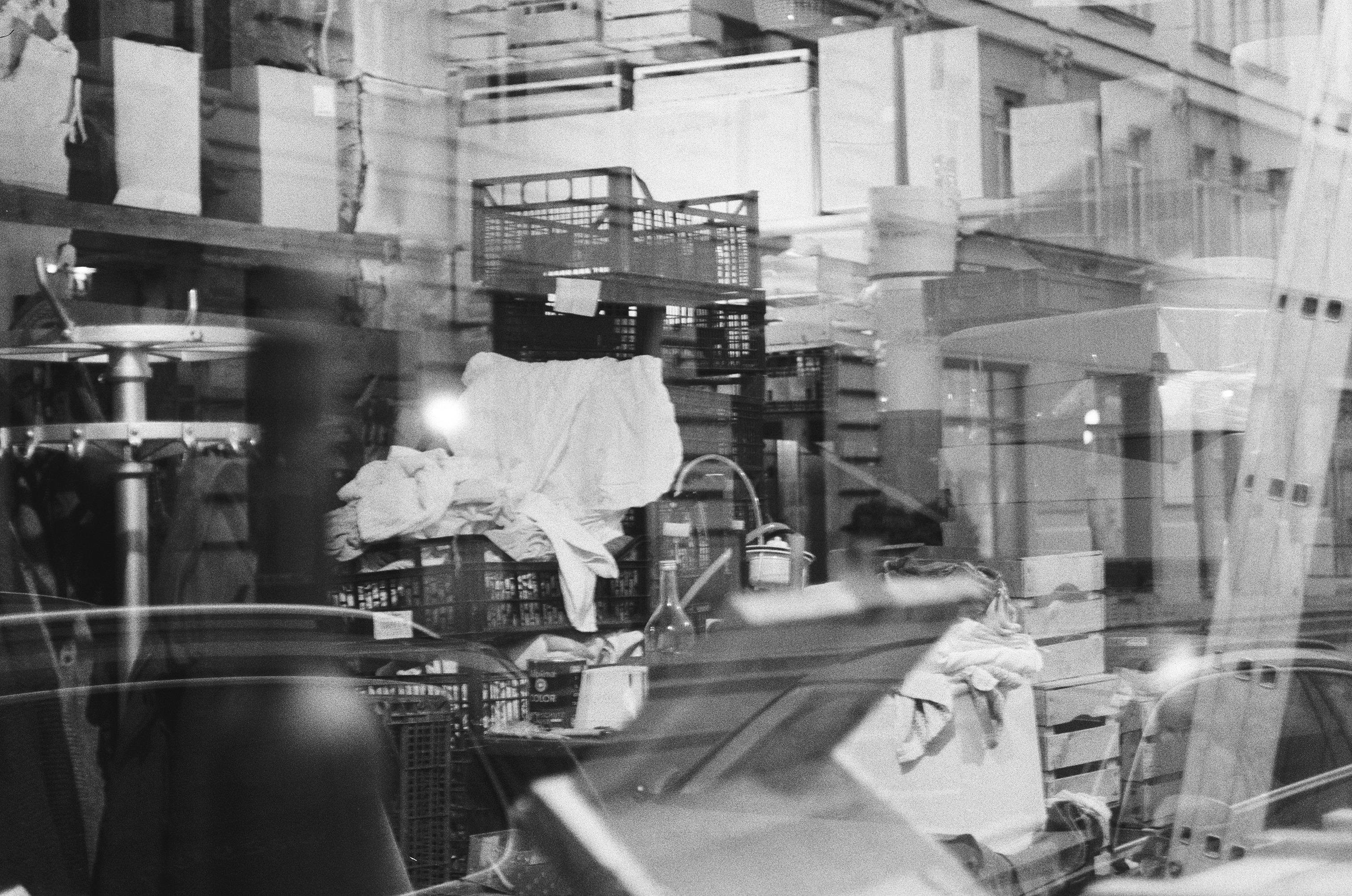 This image is a black-and-white photograph showing a cluttered storage area. There are stacks of crates or boxes, some with cloth draped over them. Various objects are scattered around, including bottles and a ladder. The scene seems to be viewed through glass, giving a reflective, layered effect, with some elements from the outside also visible.