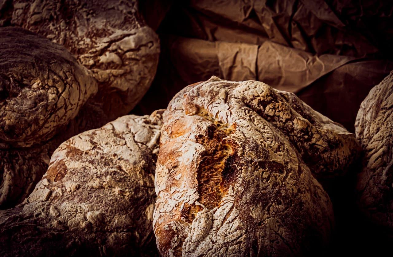 Crusty loaves of bread with a textured surface, displaying a mix of golden brown and white flour-dusted exteriors. The background consists of crumpled brown paper. The lighting highlights the rugged texture of the bread.