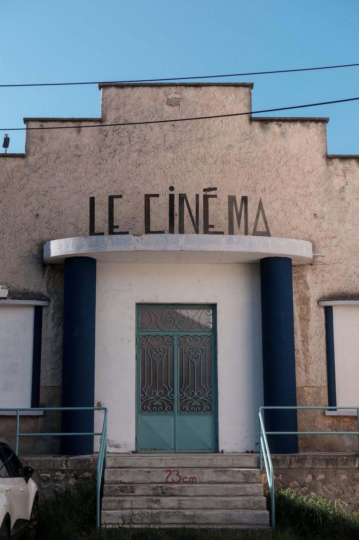 Photo verticale. Un bâtiment de face, dont le toit est en escalier, avec une entrée blanche surmontée d’une enseigne «&nbsp;Le Cinéma&nbsp;» en haut d’une volée de marches
