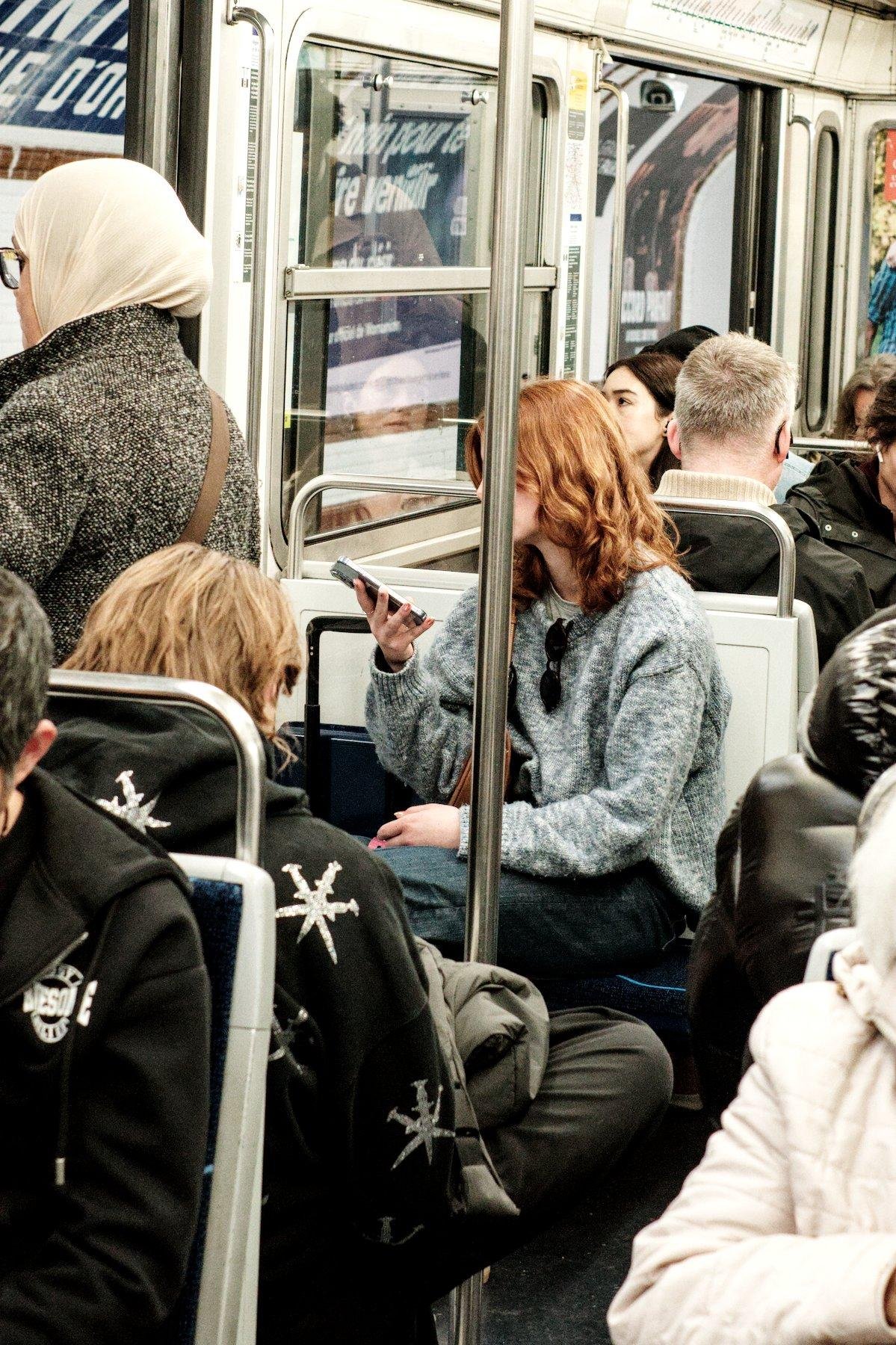 Photo verticale. Dans un wagon de métro, une jeune femme rousse aux cheveux longs aux épaules, pull gris, regarde son téléphone. Une barre verticale pour se tenir lui cache le visage, formant une séparation entre le téléphone et sa tête