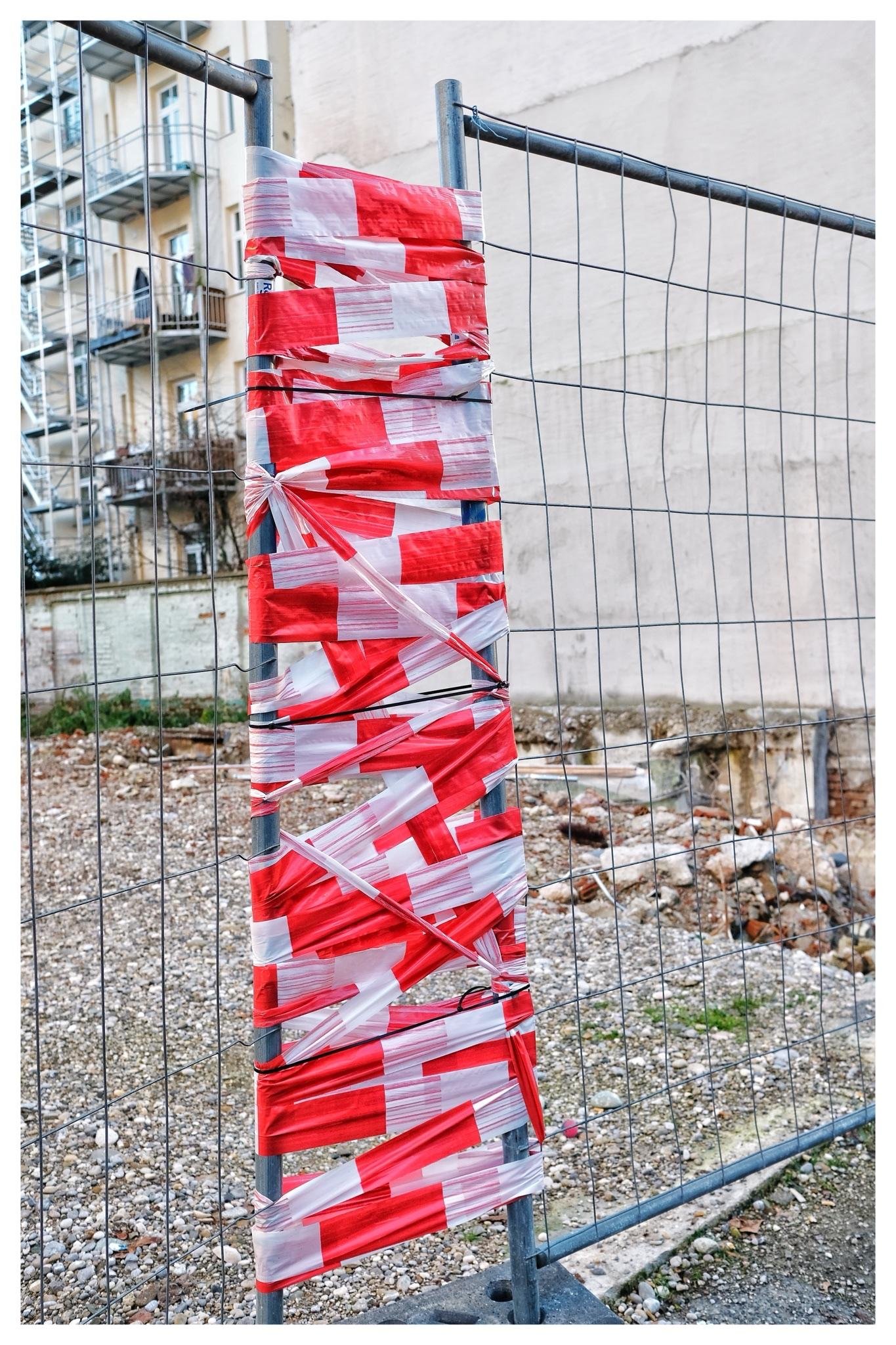 Red-and-white hazard tape wrapped around a metal fence post on a temporary wire construction fence, with a rubble-strewn vacant lot and nearby apartment building balconies in the background.