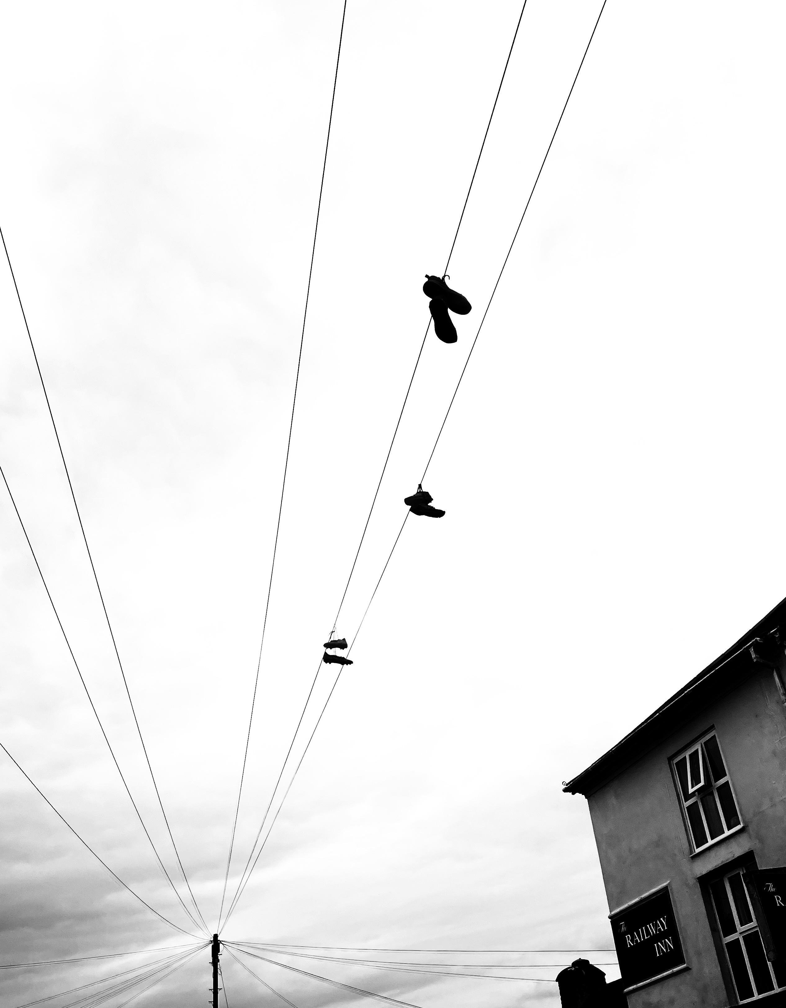 Black-and-white photo of multiple utility wires converging at a pole, with several pairs of shoes hanging from the wires; corner of a building with a “Railway Inn” sign at the lower right under an overcast sky.