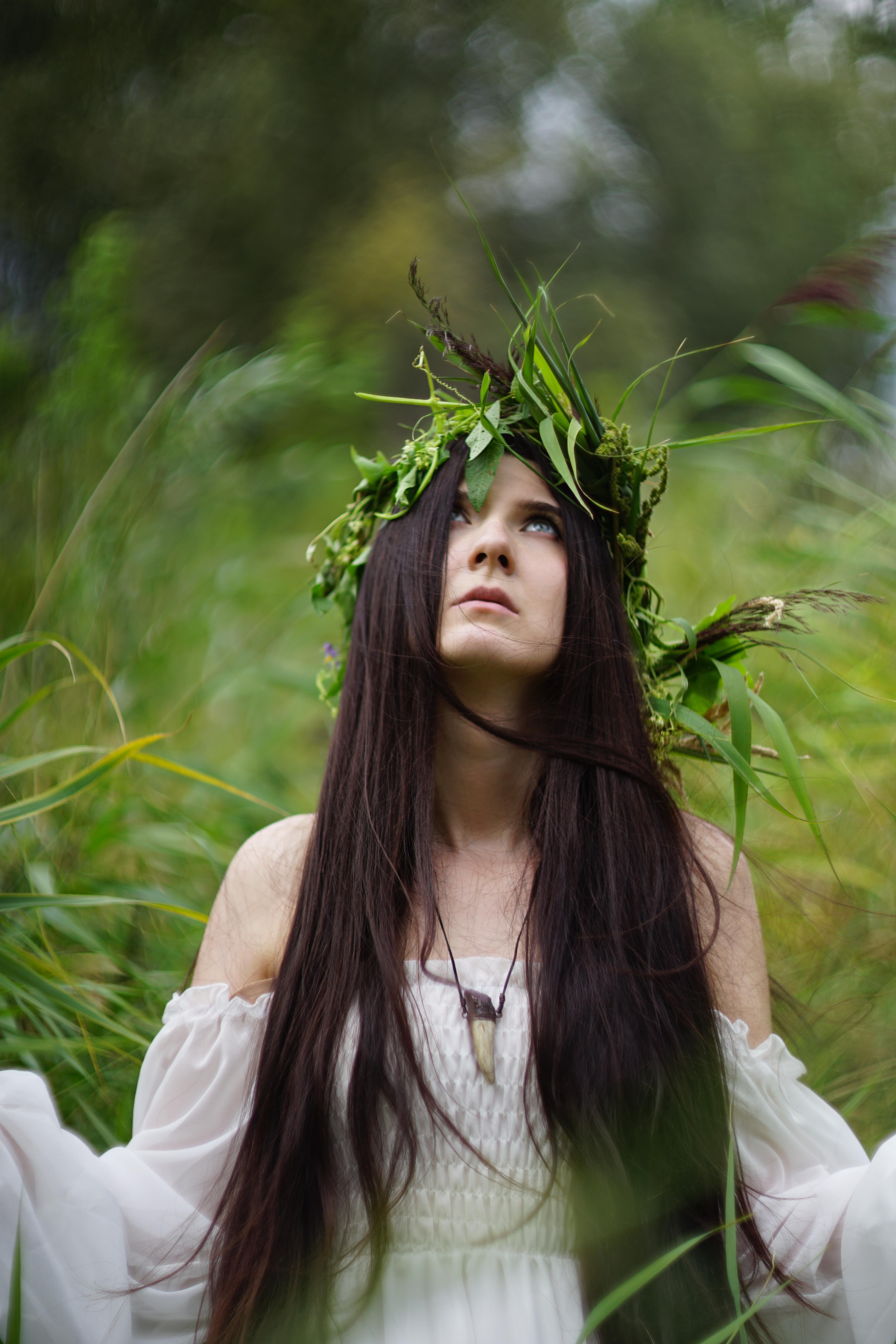 Woman with long dark hair wearing a white off-the-shoulder dress and a necklace with a tooth pendant. A crown of green leaves and grass adorns her head. She is looking upwards, surrounded by lush, blurred greenery in the background.