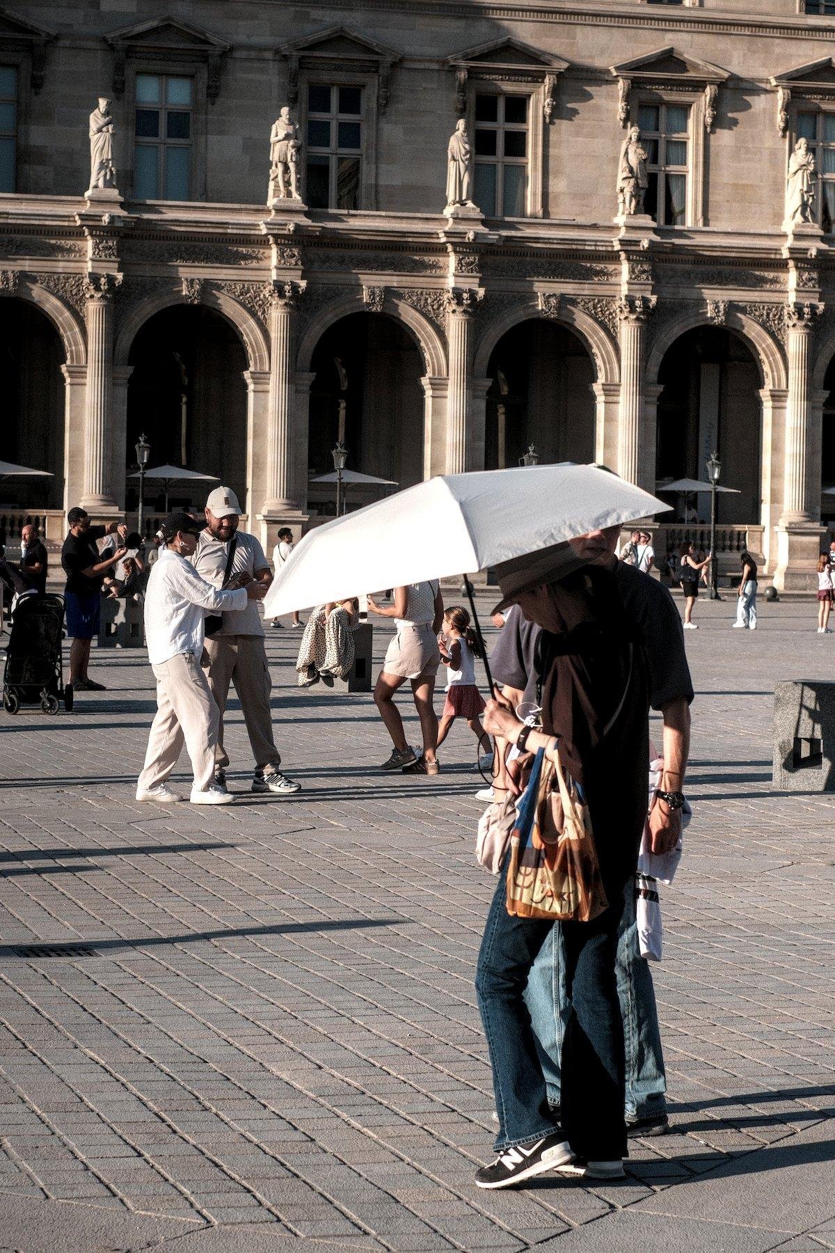 Photo verticale. Sur le parvis du Louvre, un couple consulte un téléphone à l’abri de leur ombrelle