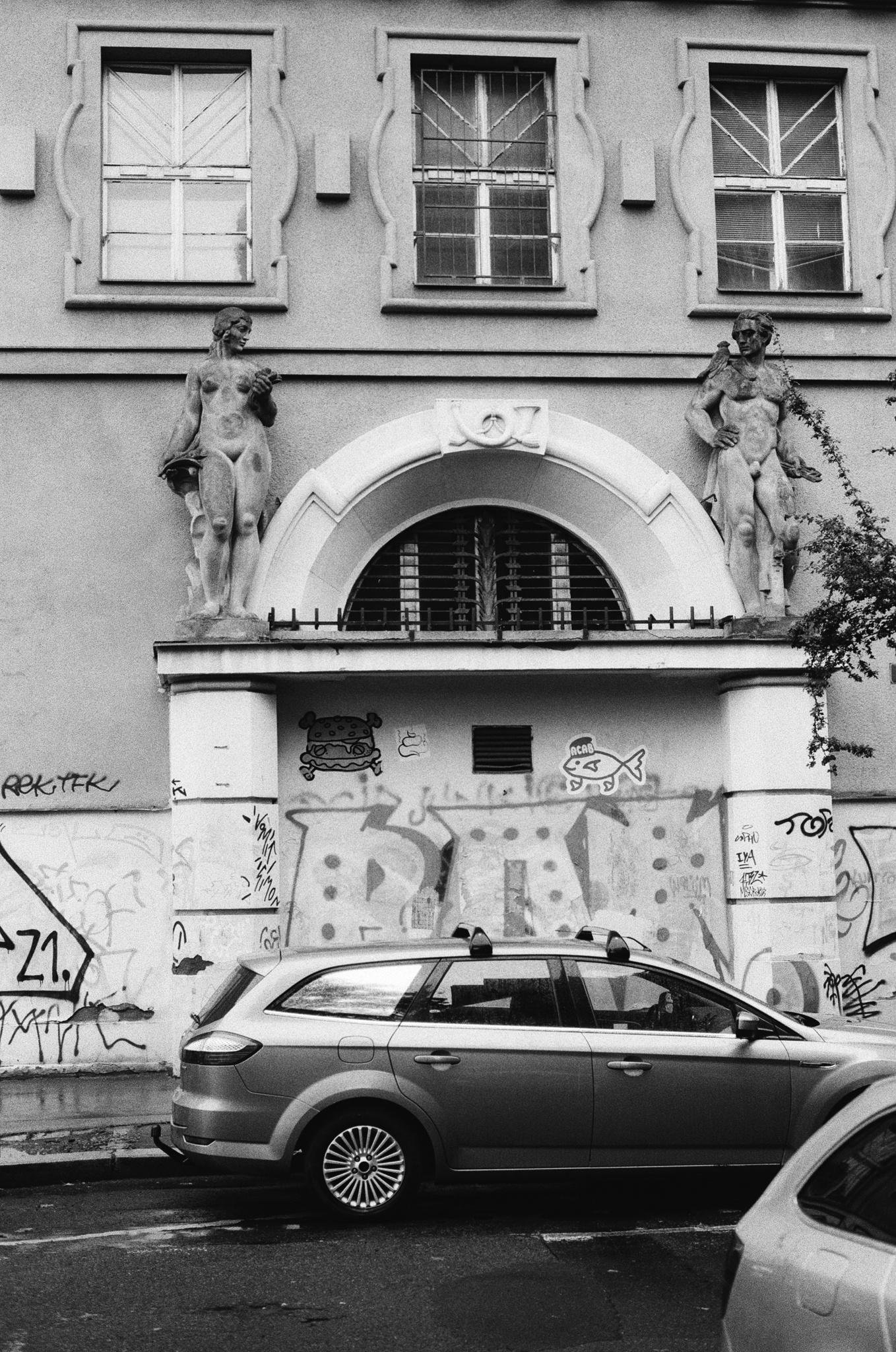 Black-and-white street view of a building facade with three upper windows and an arched entrance framed by two nude stone statues; graffiti covers the lower wall, and a station wagon is parked along the curb in front.