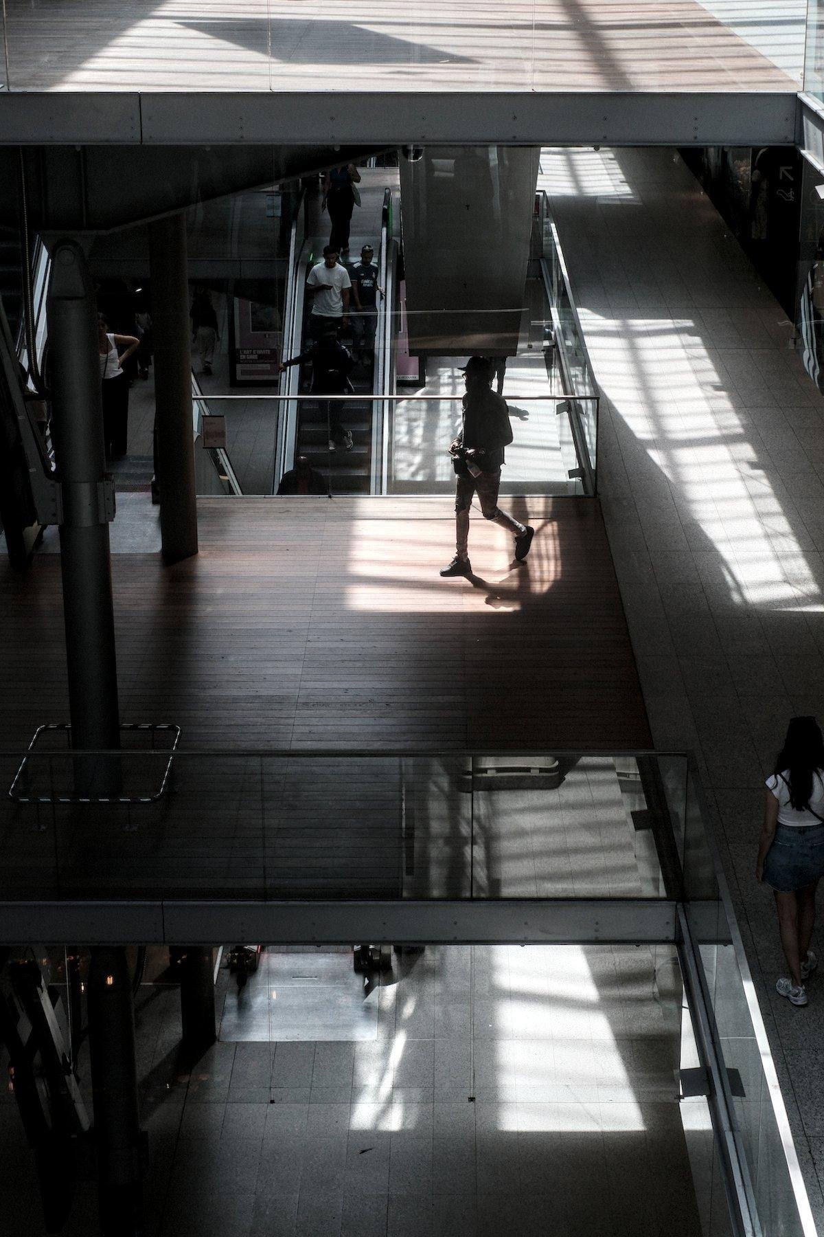 Photo verticale. Vue sur une passerelle en plancher depuis l’étage au dessus, où un carré de lumière est dessiné sur le plancher et une personne est en train de le traverser. Derrière, un escalator avec des gens dans le sens descendant
