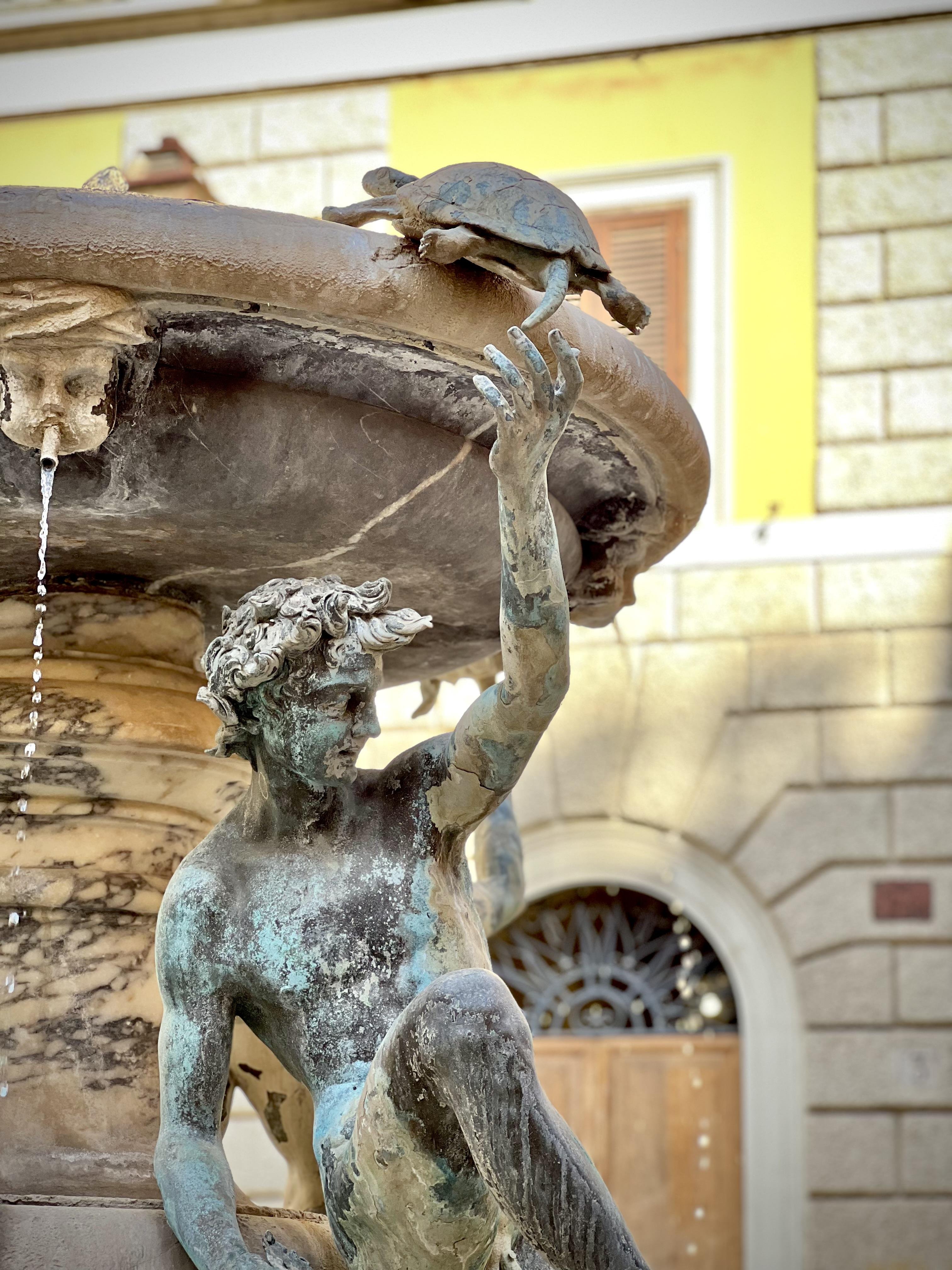 Vista dettagliata di una fontana. C'è una scultura di una figura maschile, possibilmente un personaggio mitologico, che si protende verso l'alto. Sul bordo della fontana c'è una tartaruga. L'acqua scorre dalla struttura sopra la fontana. Sullo sfondo si vede parte di un edificio con una finestra e decorazioni in pietra.