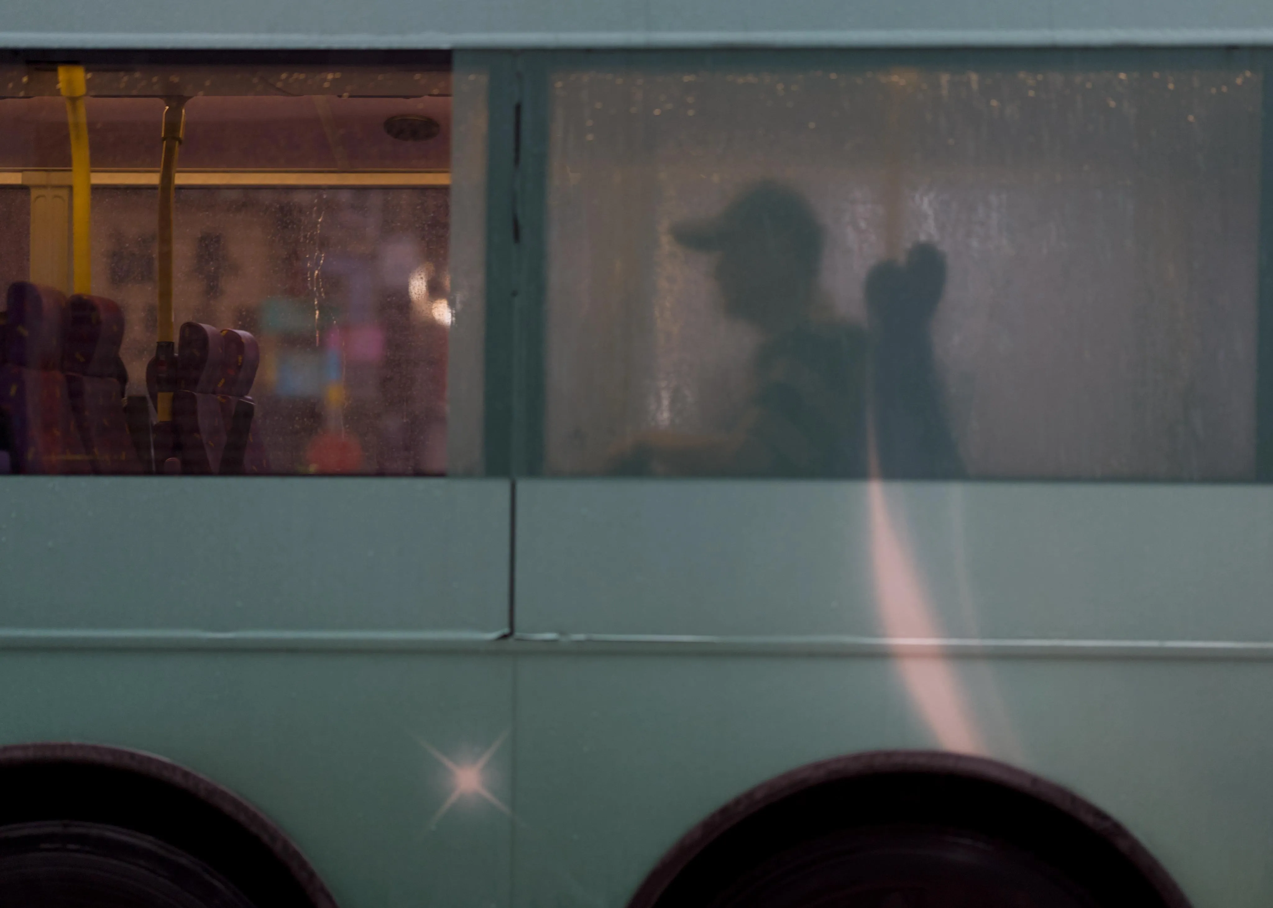 Photograph of a bus from its side, on a rainy day. The body color of the bus is teal. In the left half of the picture, the bus window is see-through and empty seats are visible through the window. In the right half of the picture, the bus window is translucid but more opaque and the silhouette of a man with a baseball cap sitting by the window is visible.