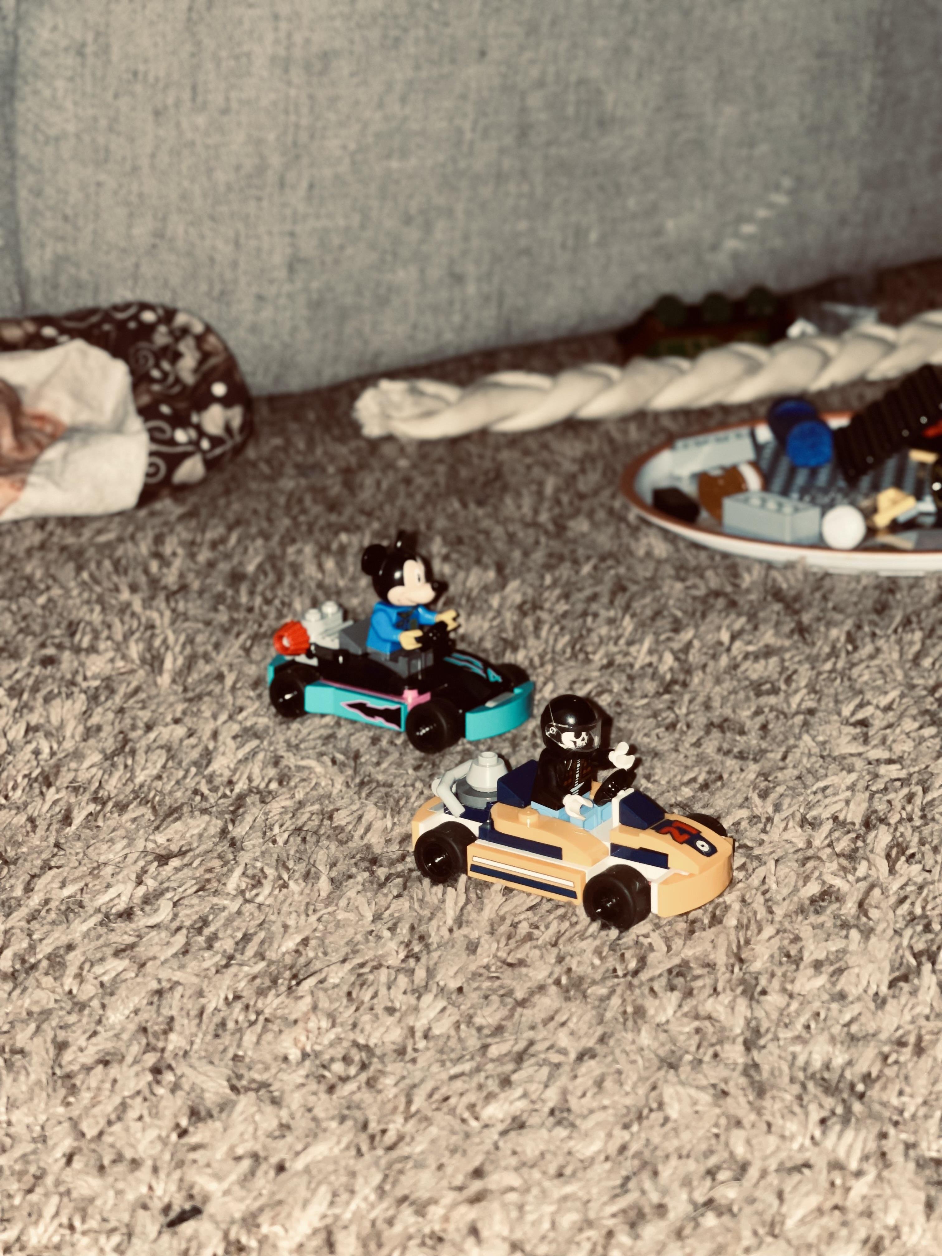 The image shows two LEGO-style toy vehicles with miniature figures inside. The figures appear to be characters, and they're set on a carpeted floor. In the background, there's a rope toy and a collection of more LEGO pieces on a plate.