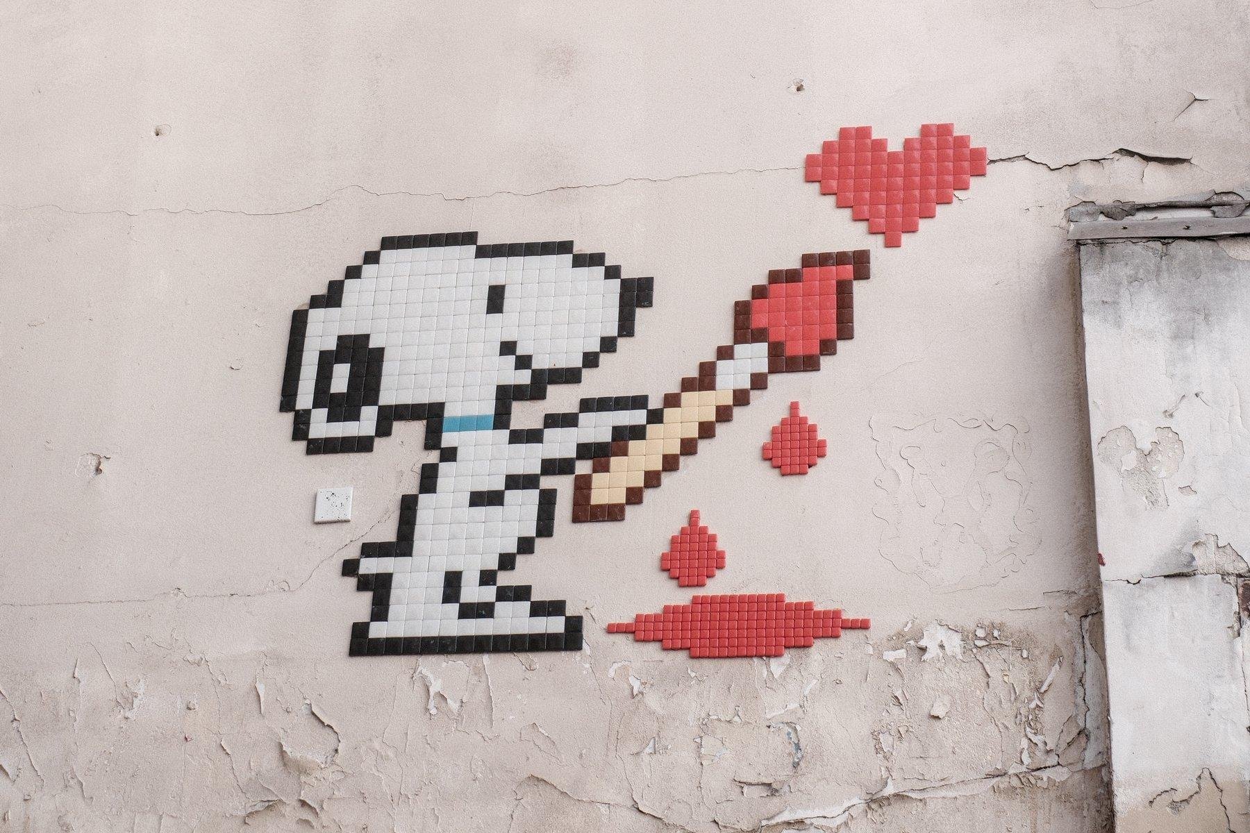 Photo horizontale. Pixelart sur un mur de Snoopy tenant un pinceau duquel goutte de la peinture rouge en une flaque, avec lequel il a peint un cœur rouge