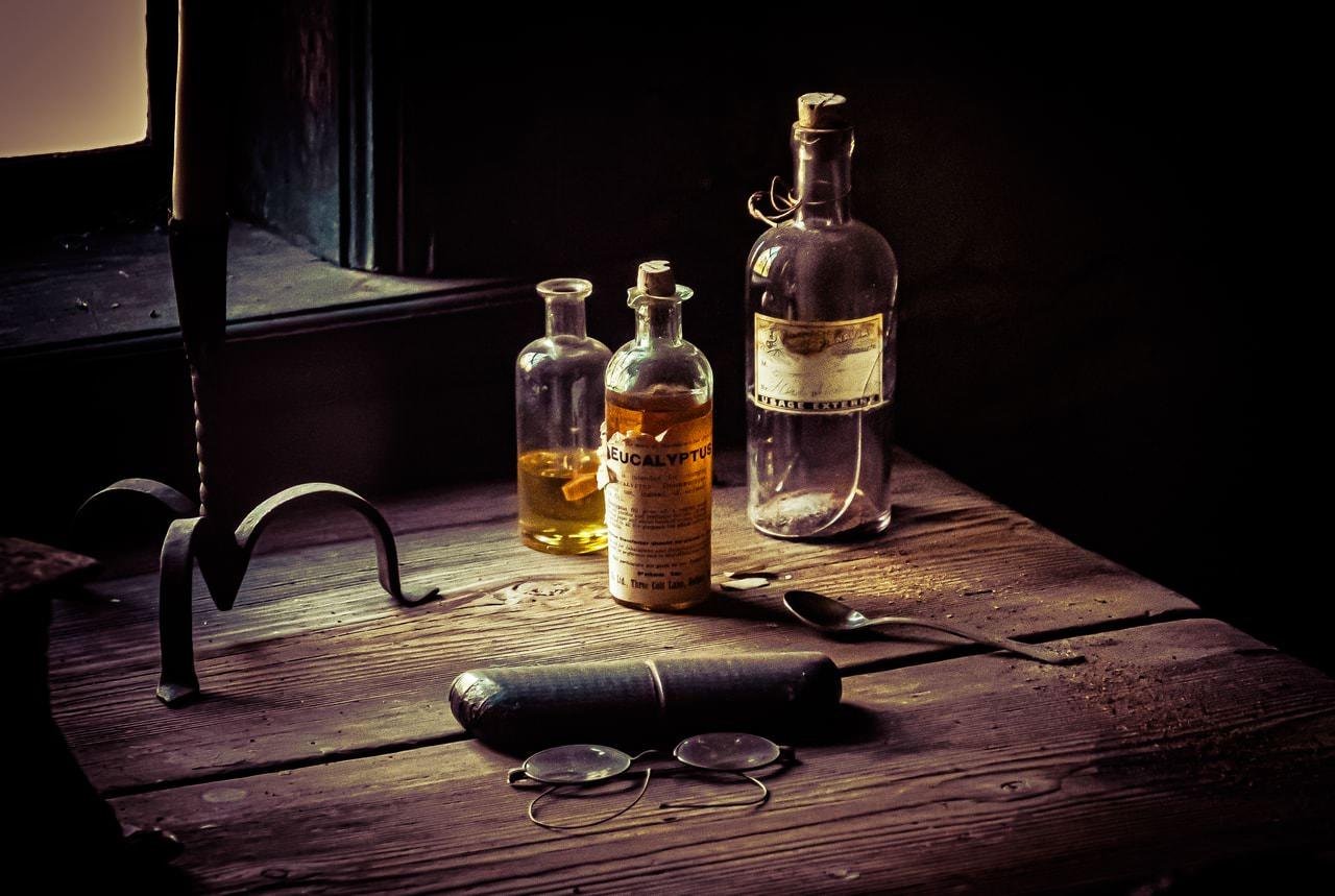 Old wooden table with three vintage glass bottles, one labeled "Eucalyptus," and a tarnished spoon beside them. An unlit candle stands next to the bottles. A pair of round spectacles and a glasses case rest in the foreground. Dim lighting creates shadows, adding a rustic atmosphere.