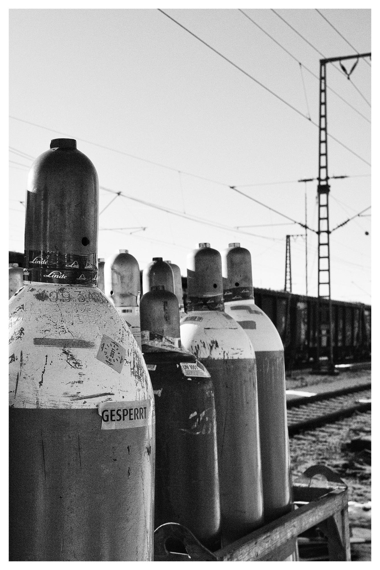 Black and white photograph of several large, industrial gas cylinders standing upright on a wooden pallet. The cylinders are weathered and have labels, one of which reads "GESPERRT." Overhead power lines and a steel tower are visible in the background, indicating an outdoor setting, possibly near a railway. The image captures the rugged texture and industrial environment.