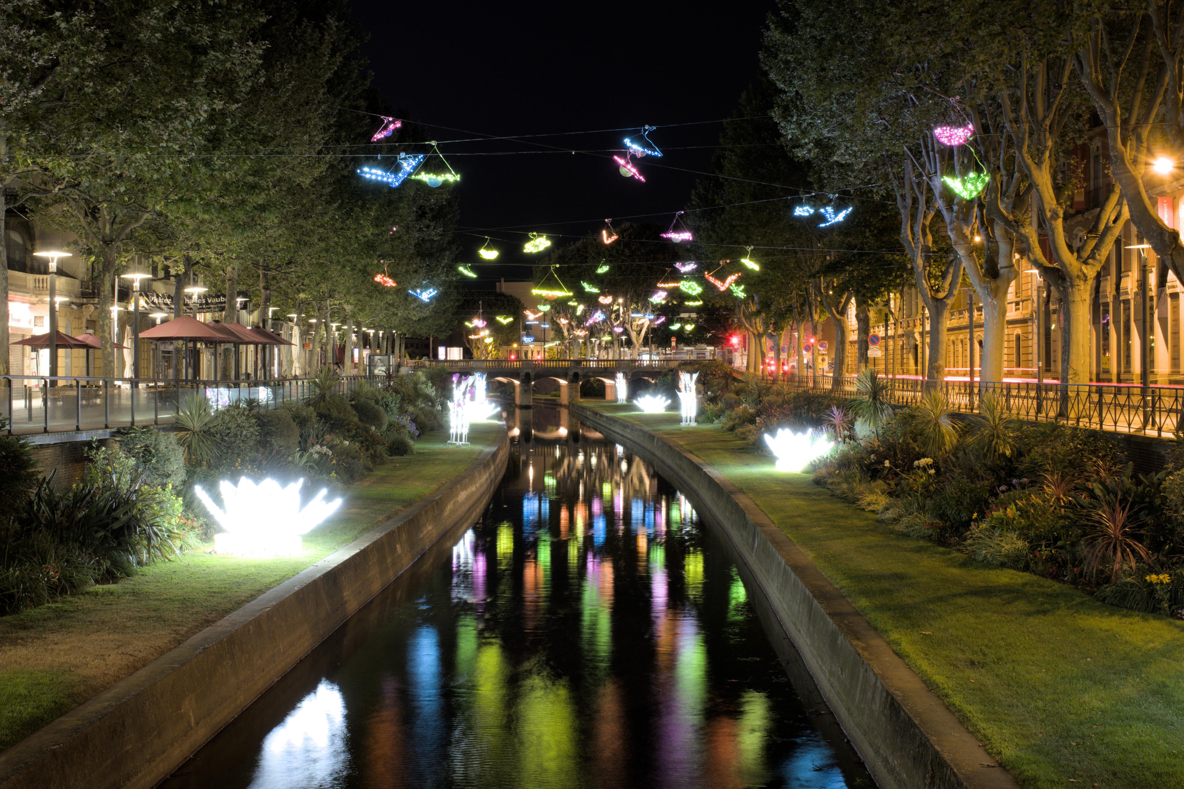 This image shows a canal or river at night, lined with trees and illuminated with colorful lights hanging above the water. Both sides of the canal are well-lit, and the reflections of the lights can be seen on the water. There are also bright, flower-shaped lights on the grassy banks, adding to the festive atmosphere.