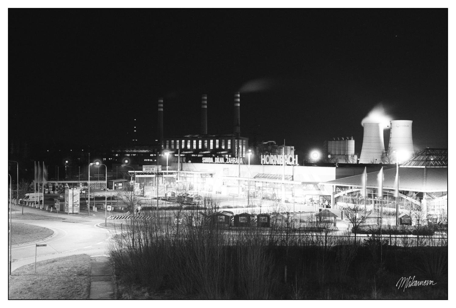 The image is a black and white nighttime photograph of an industrial area. In the foreground, there's a lighted building with the name "HORNBACH" visible, which suggests it's a large retail store or warehouse. In the background, there are industrial structures such as smokestacks and cooling towers emitting steam or smoke, indicating a factory or power plant. The scene includes streetlights and a mostly empty road. The overall atmosphere is industrial and urban.