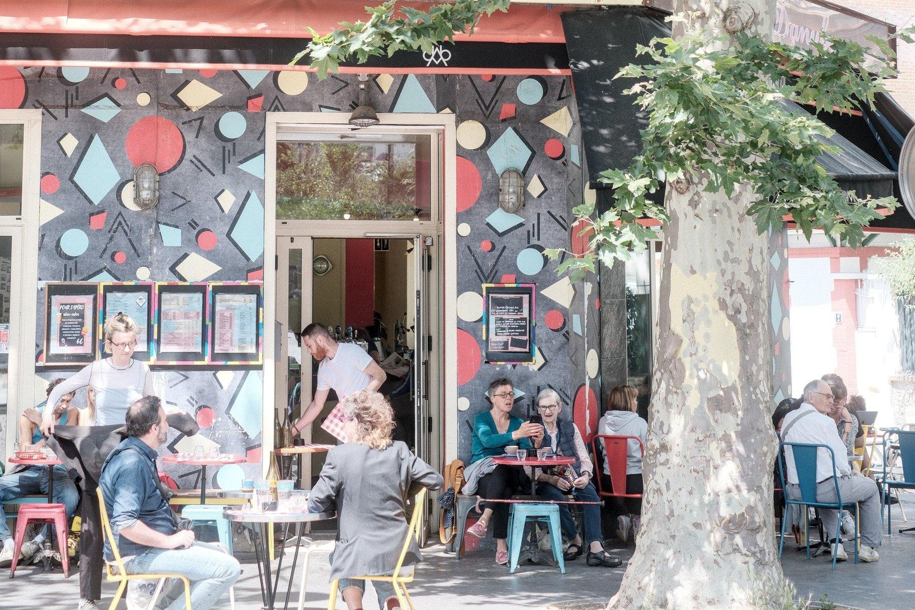 Photo horizontale. Une terrasse de café avec des gens dont la façade est grise avec des disques et losanges bleu, rouge ou blanc, me faisant penser à un générique de série des années 80
