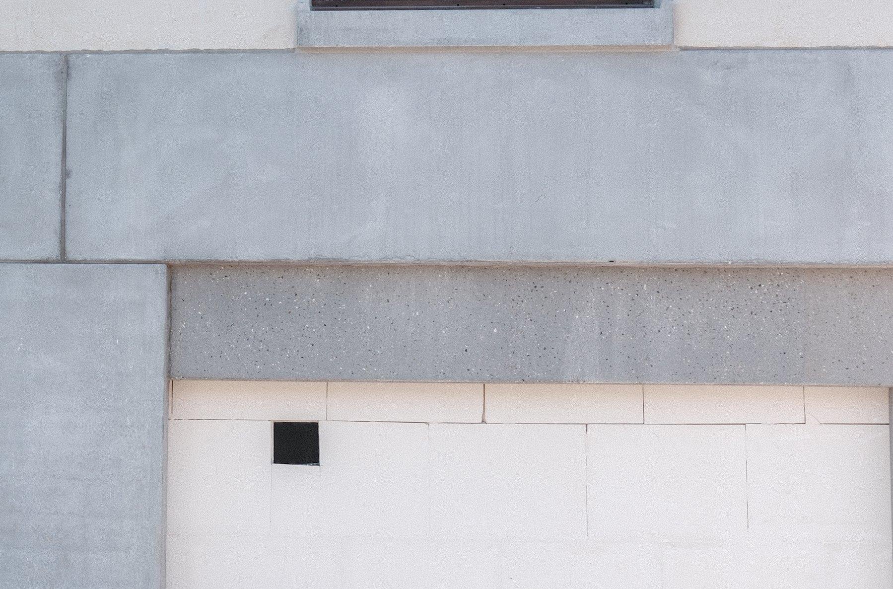 Photo horizontale. Un mur en briques blanches, non peint encadré par des blocs de béton gris, dans lequel il y a un petit trou carré