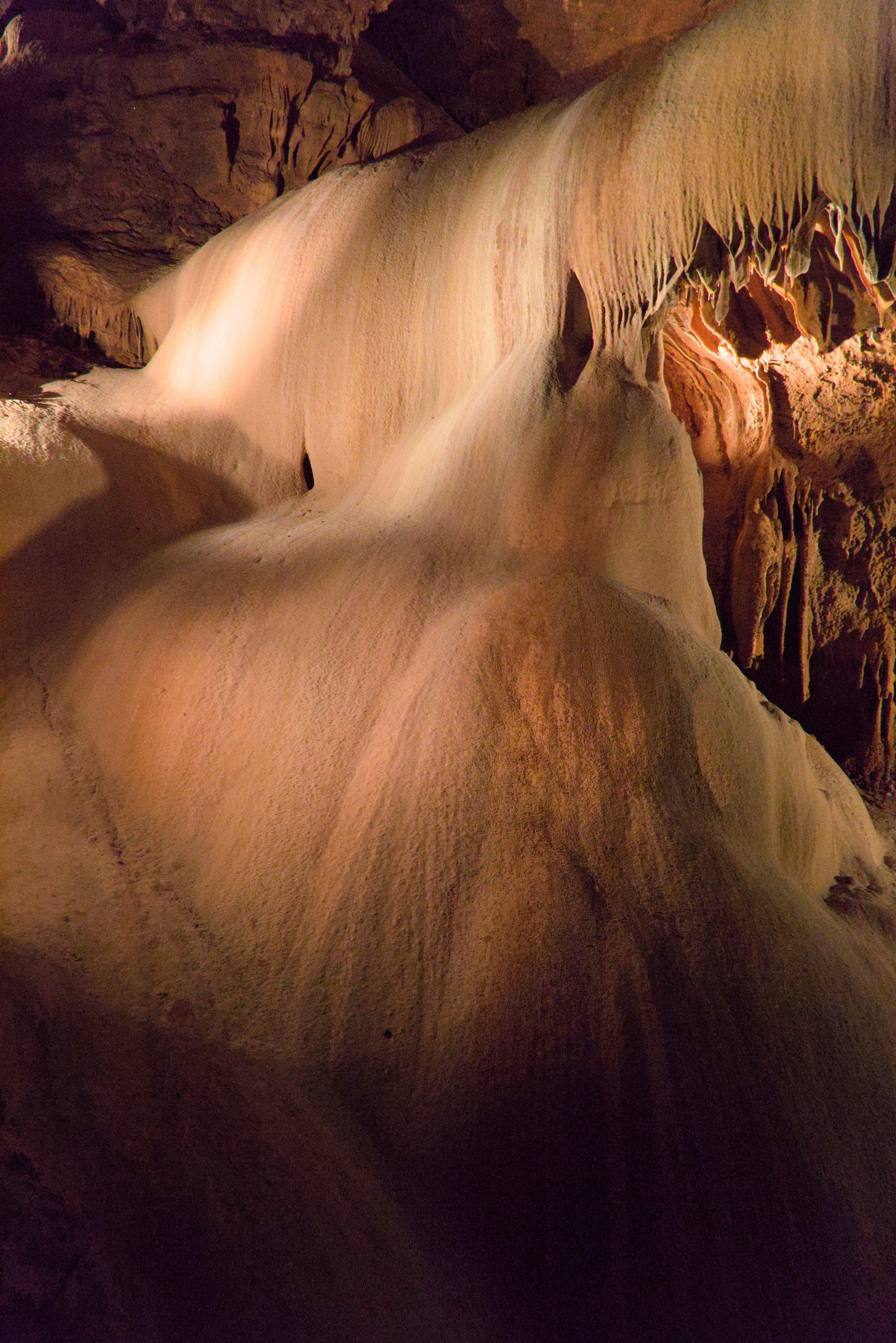 Photo verticale. Une formation rocheuse, sous terre, faisant penser à une cascade d’eau pétrifiée, éclairée avec des lumières orangées