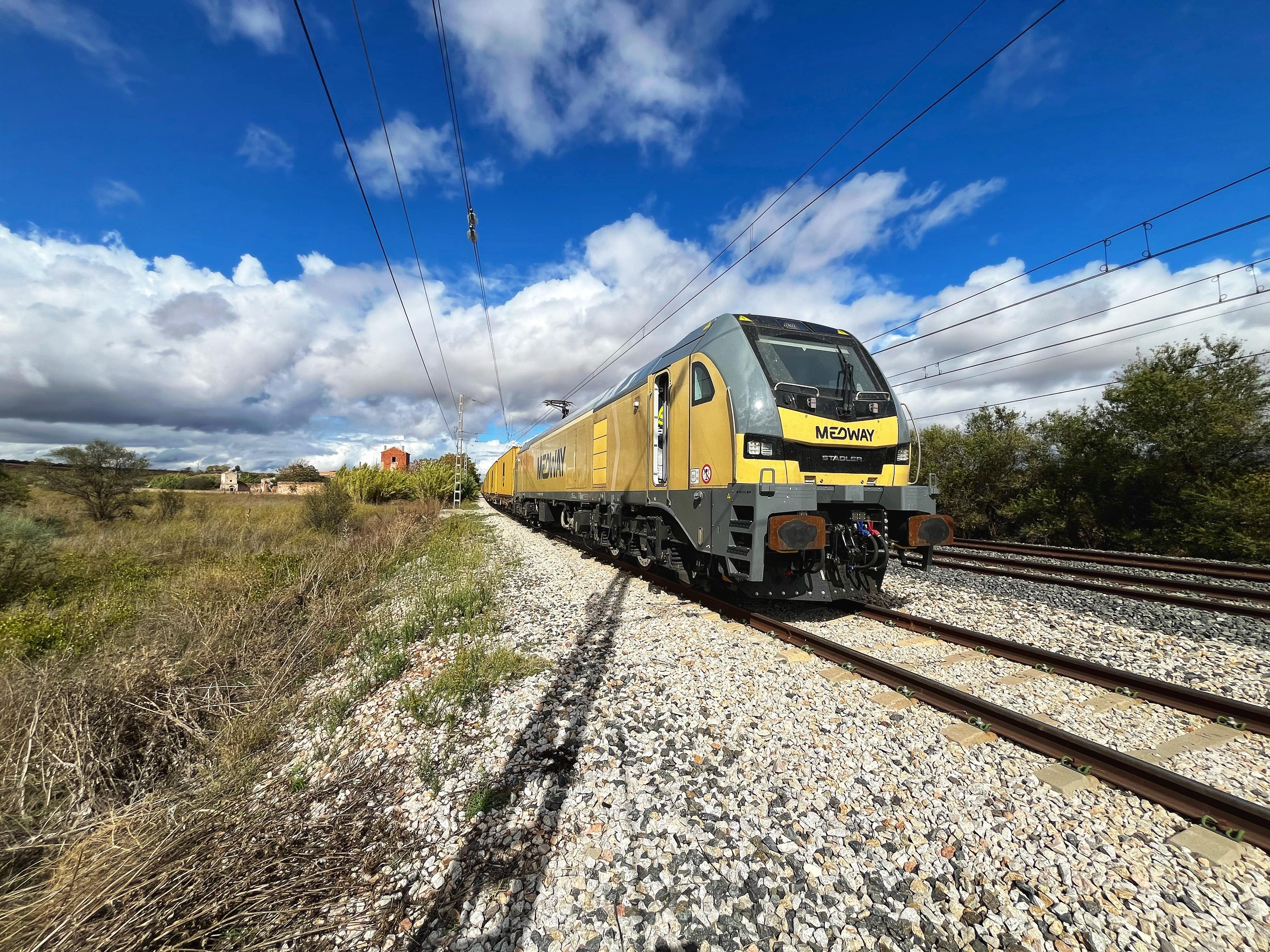 The image shows a freight train traveling on a railway track. The train has a gray and yellow locomotive, and appears to be in a rural setting with grass and bushes nearby. The sky is partly cloudy with a mix of blue sky and white clouds. The word "MEDWAY" is visible on the train.