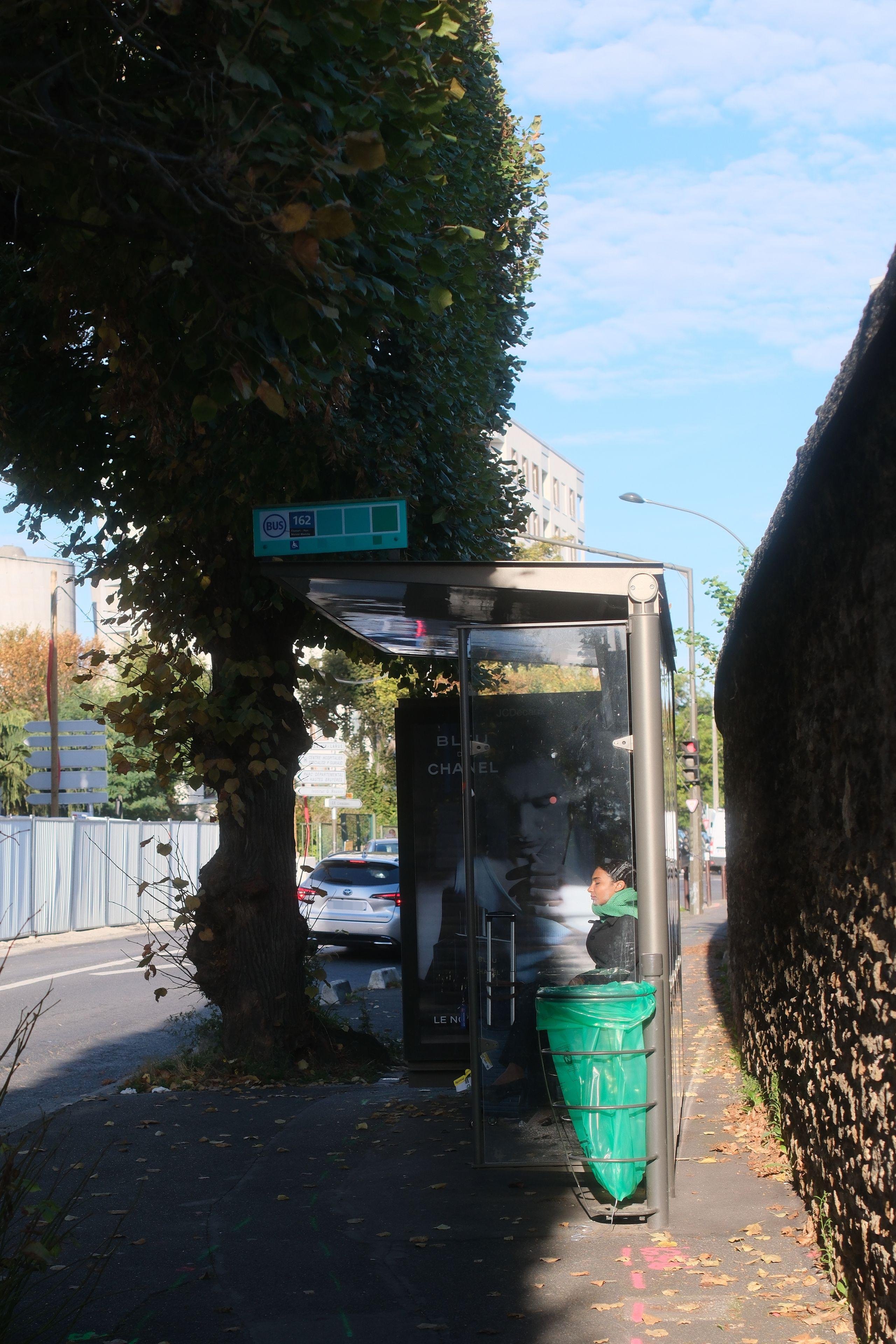 Photo verticale. Un arrêt de bus où est assise une jeune femme, éclairée par un rayon de soleil alors que le reste de l’abris est dans l’ombre. Elle a les yeux fermés, savourant la chaleur du soleil
