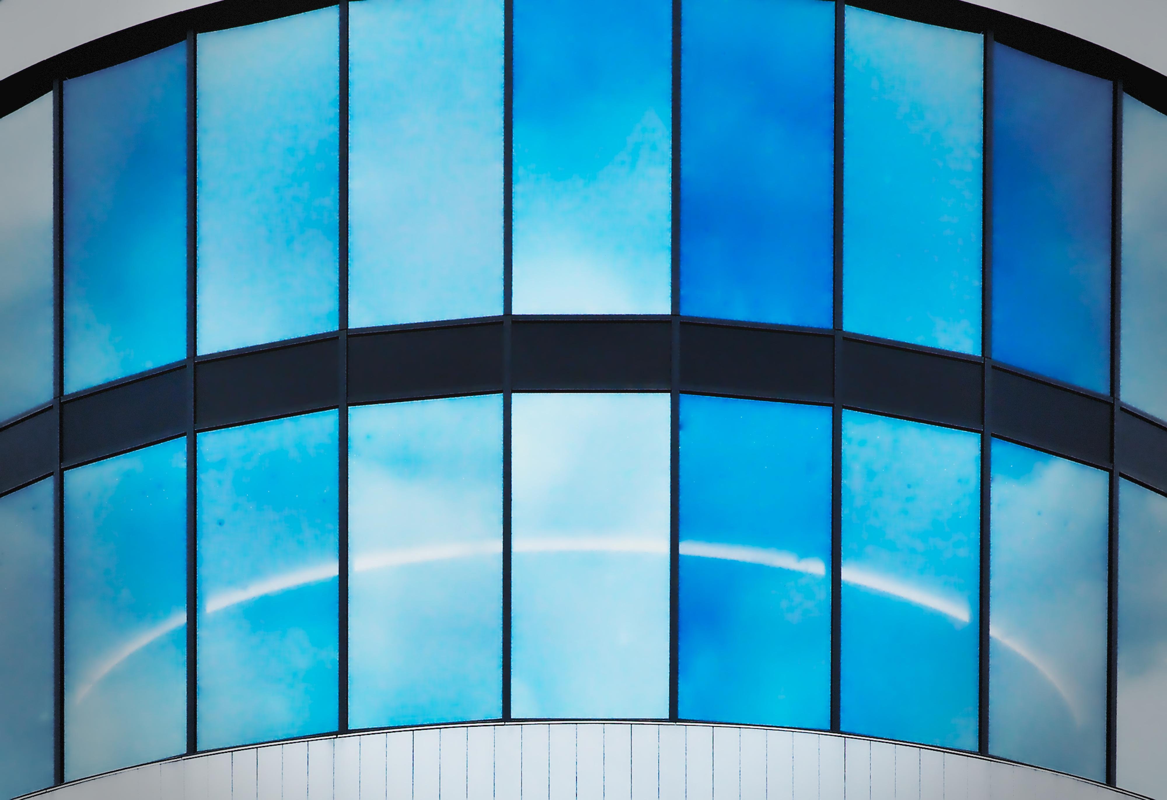 Curved glass facade composed of large, vertical panels reflecting a bright blue sky with scattered white clouds. Horizontal black bands divide the panels. The glass creates a rainbow-like arc across the center, enhancing the architectural design. 
### 
Die geschwungene Glasfassade besteht aus großen, vertikalen Paneelen, die einen strahlend blauen Himmel mit vereinzelten weißen Wolken spiegeln. Horizontale schwarze Streifen unterteilen die Paneele. Das Glas bildet einen regenbogenartigen Bogen in der Mitte und unterstreicht so das architektonische Design. 