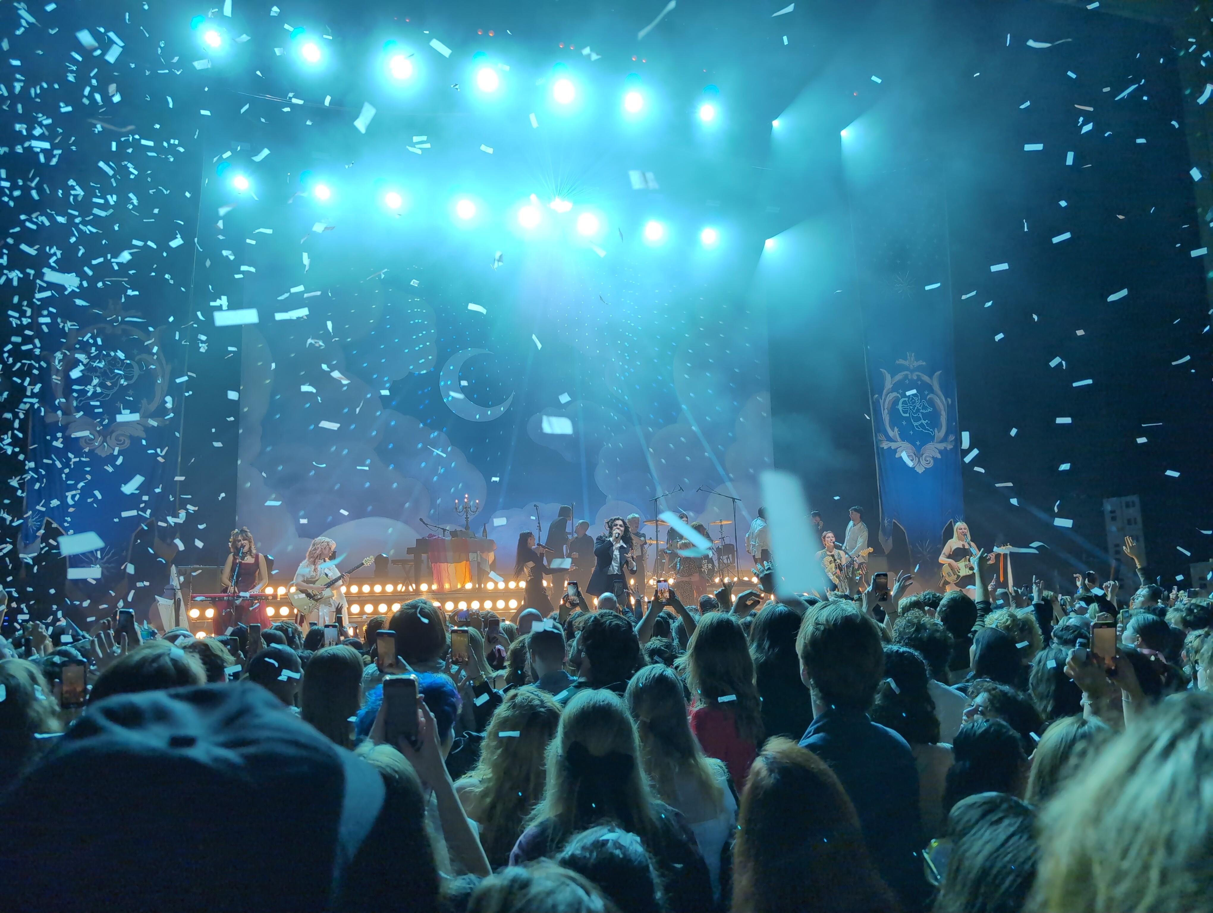 This image shows a concert scene with a band performing on stage. There are bright lights overhead, and confetti is falling over the crowd. The audience is engaged, with many people holding up their phones, possibly recording or taking photos of the performance.