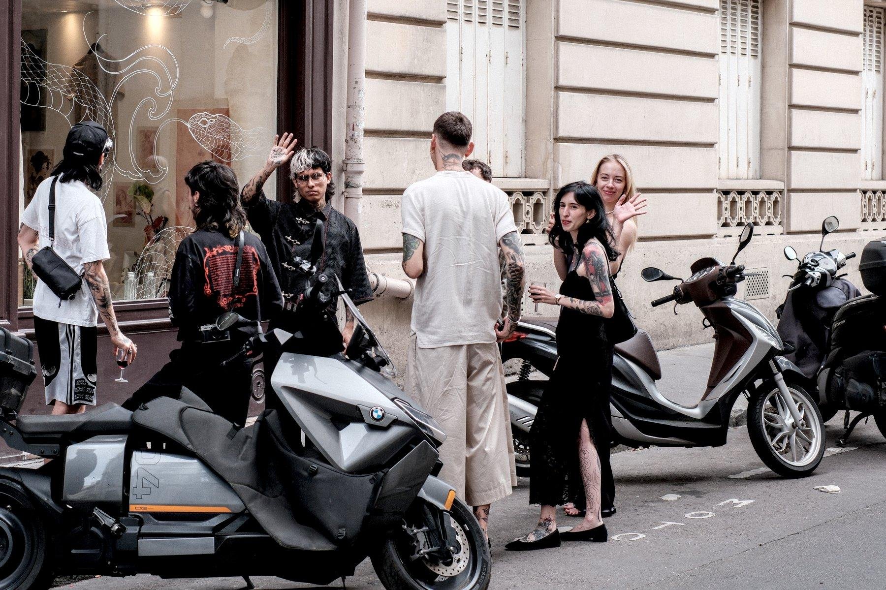 Photo horizontale. Dans une rue, entre les scooters garé à un emplacement réservés aux motos, des gens au look divers (cheveux bicolores, tatouages, robe, chemise de soirée…) font une pause cigarette et m’ont remarqué. L’un d’eux me fait signe de la main