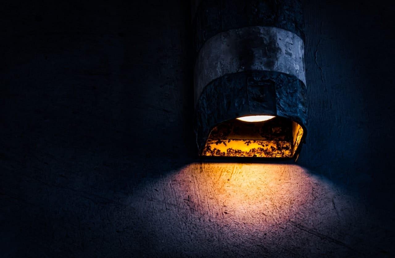 Wall-mounted outdoor light fixture with a weathered metal hood, casting a warm yellow beam onto a dark textured wall at night.