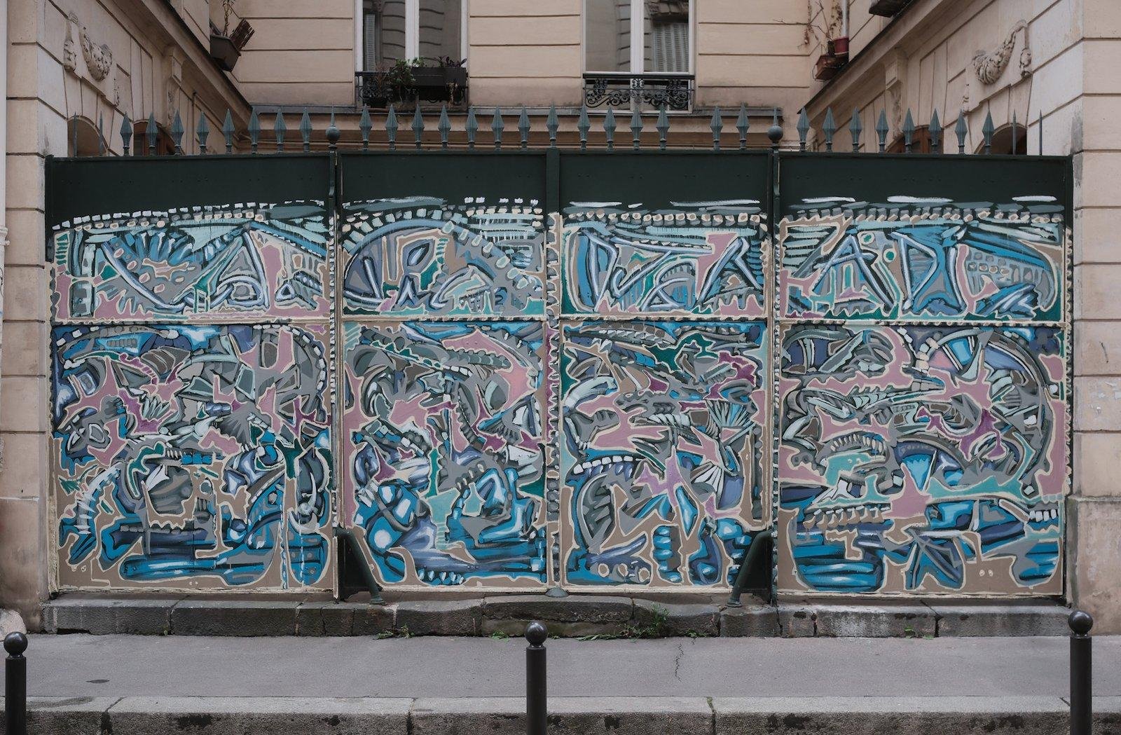 Photo format portrait montrant un mur métallique fermant une cours d’immeuble  presque entièrement couverte de streetart abstrait dans une palette de couleurs homogène bleus roses blanc