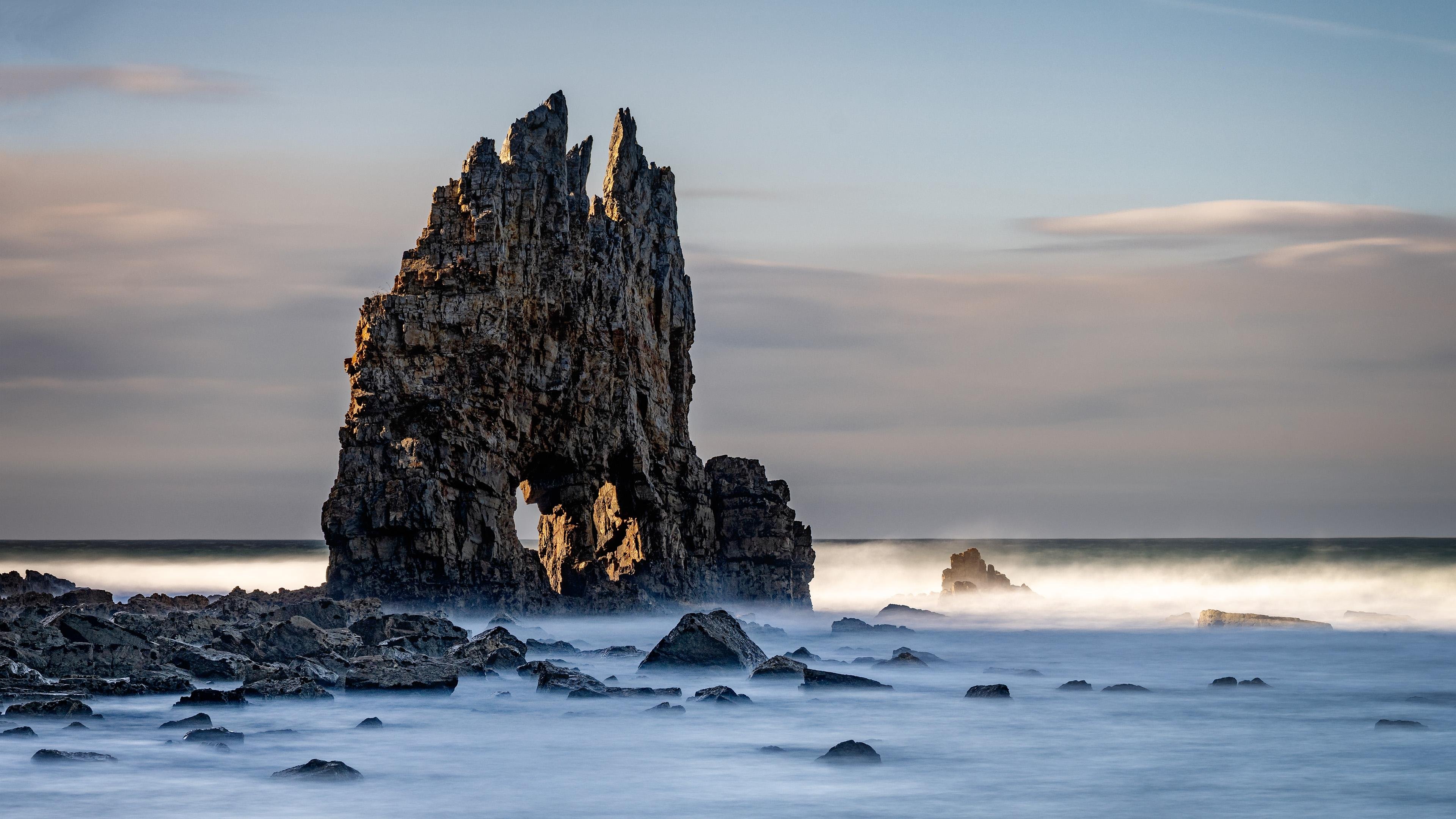 Rock formation rising from the sea with jagged peaks, illuminated by soft sunlight. Surrounding the rock are calm, misty waters and scattered smaller rocks. In the background, a cloudy sky meets the horizon, creating a serene coastal scene.