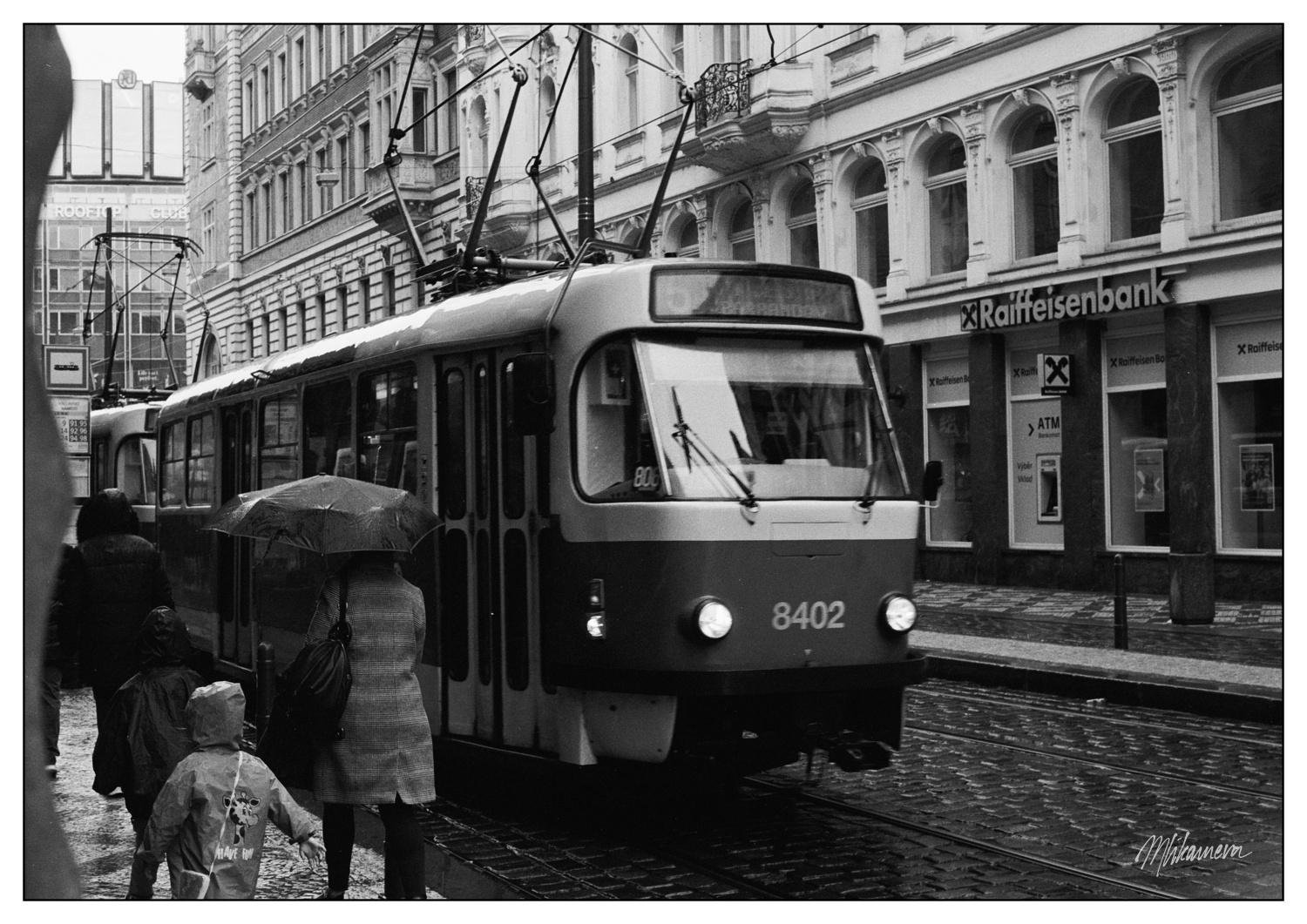 A monochrome photograph of a street where a person under an umbrella and a child walk away from the viewer while a tram is aproaching the viewer. The set is within interwar looking buildings.
