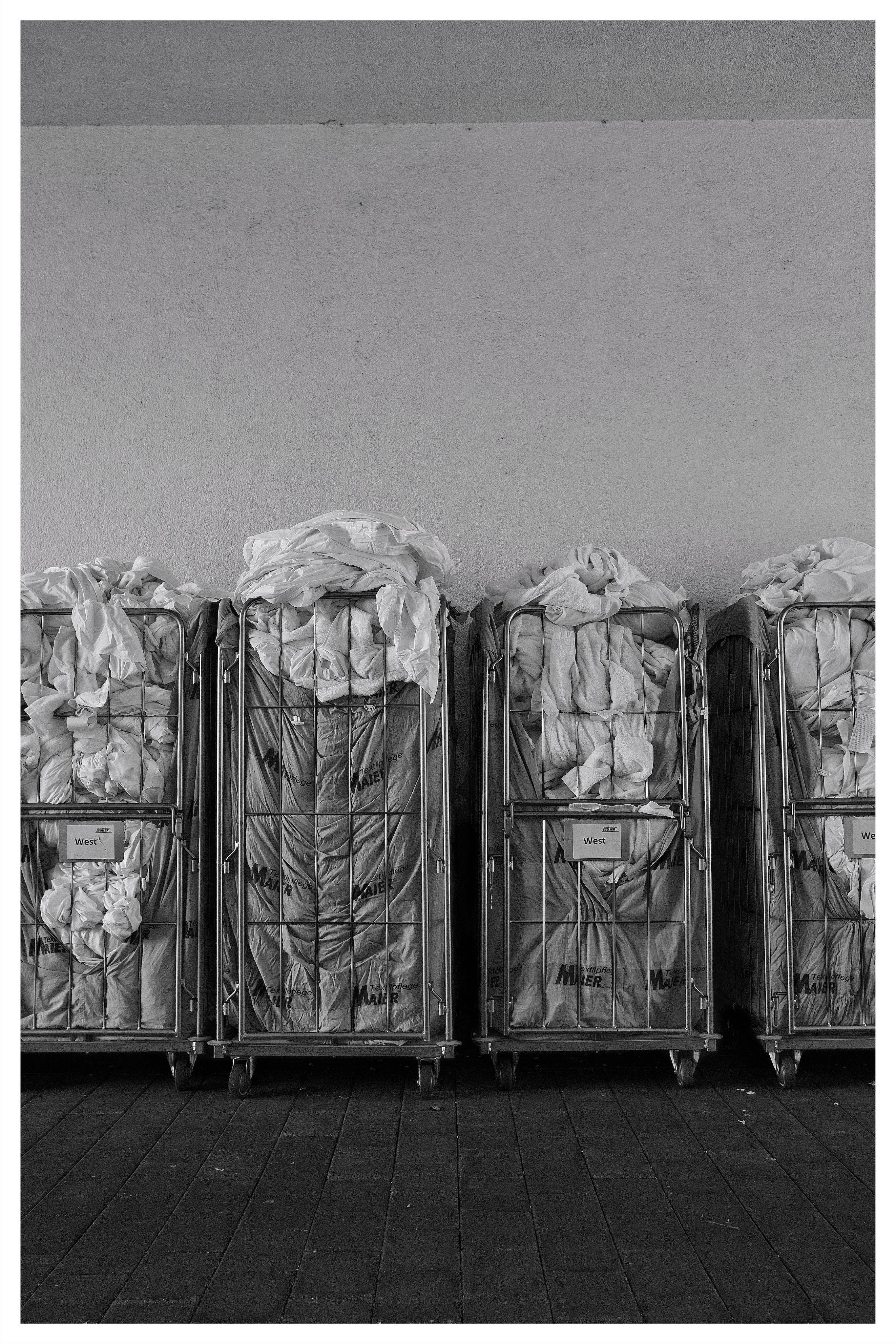 Four wheeled metal laundry carts lined up against a plain wall, each stuffed with rumpled white linens and covered with mesh or fabric panels; dark tiled floor in the foreground.