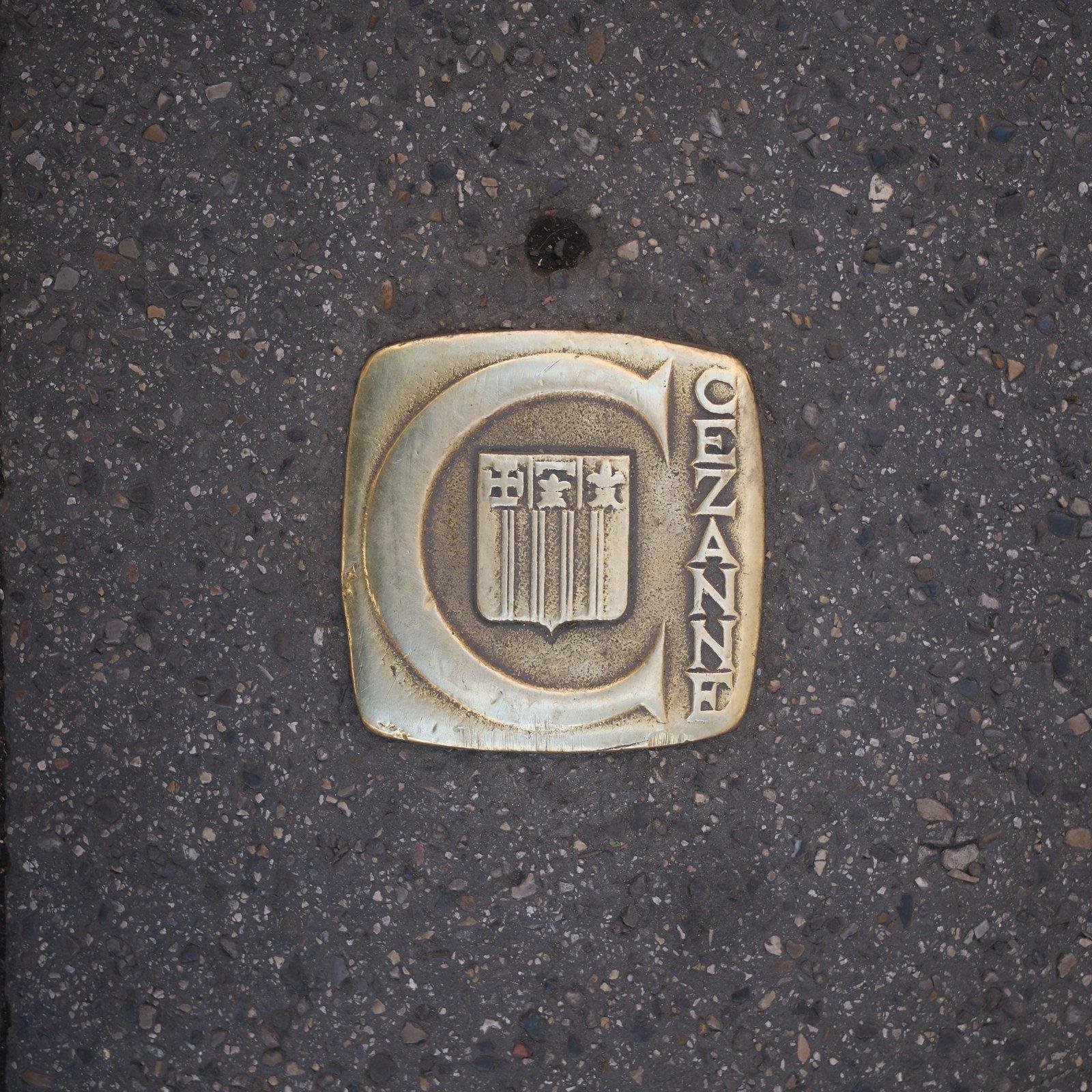 Photo carrée. Un écusson doré Cezanne incrusté dans le bitume d’un trottoir, vu de dessus