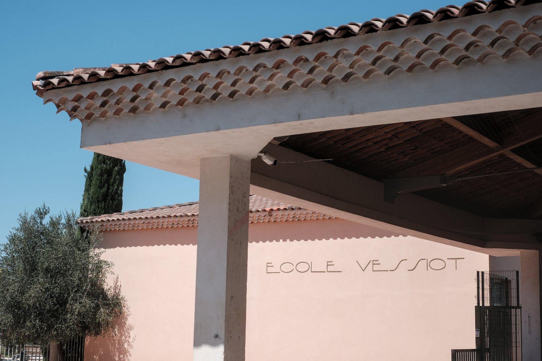 Photo horizontale. Un bout de toit de préau d’une école, et en dessous, en arrière plan, le nom de l’école « ECOLE VESSIOT » dans une police très épurée et moderne, le tout sous un ciel bleu. Un arbuste est sur le bord gauche de l’image et un bout de cyprès joint l’arrière plan et le premier plan avec le toit