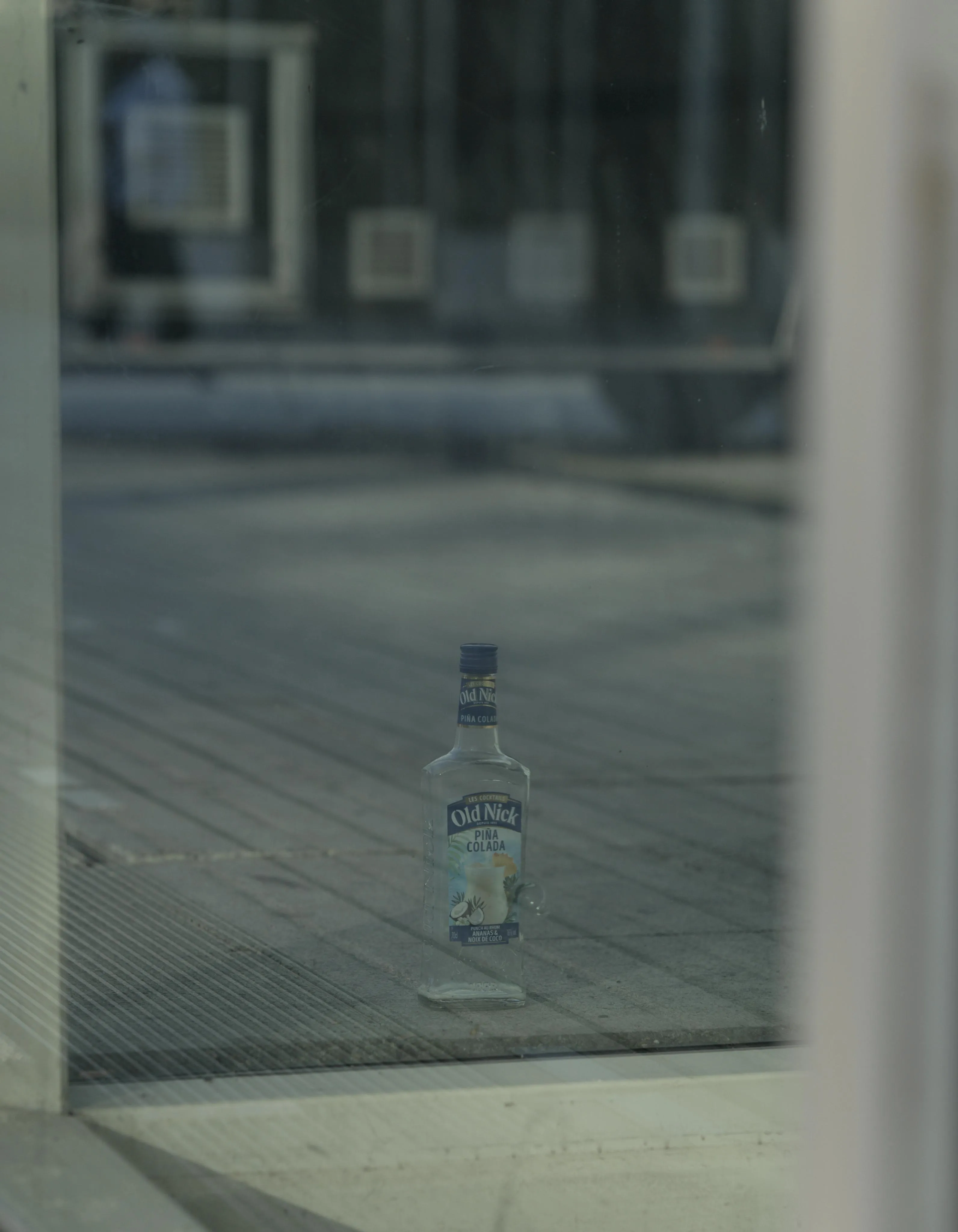 Photograph of an empty glass bottle of alcoholic Pina Colada laid on the ground, seen from behind the glass of a window. The bottle has a deep blue label including an illustration of a glass of pina colada. The ground on the other side is reflected in the window. Further back, the wheels and bottom of the body of a truck can be seen.