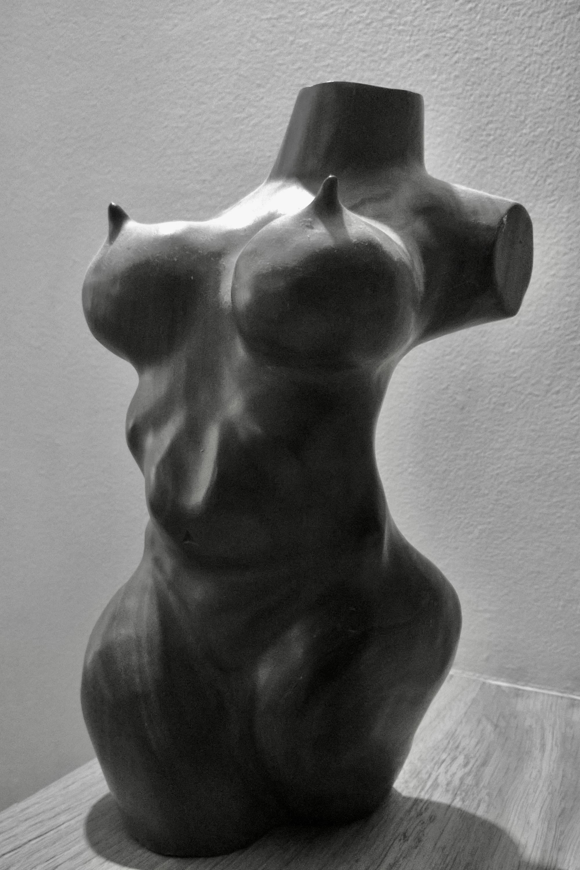 Black-and-white photo of a smooth, headless female torso sculpture with one arm missing and the other cut at the shoulder, positioned on a wooden surface against a plain wall.