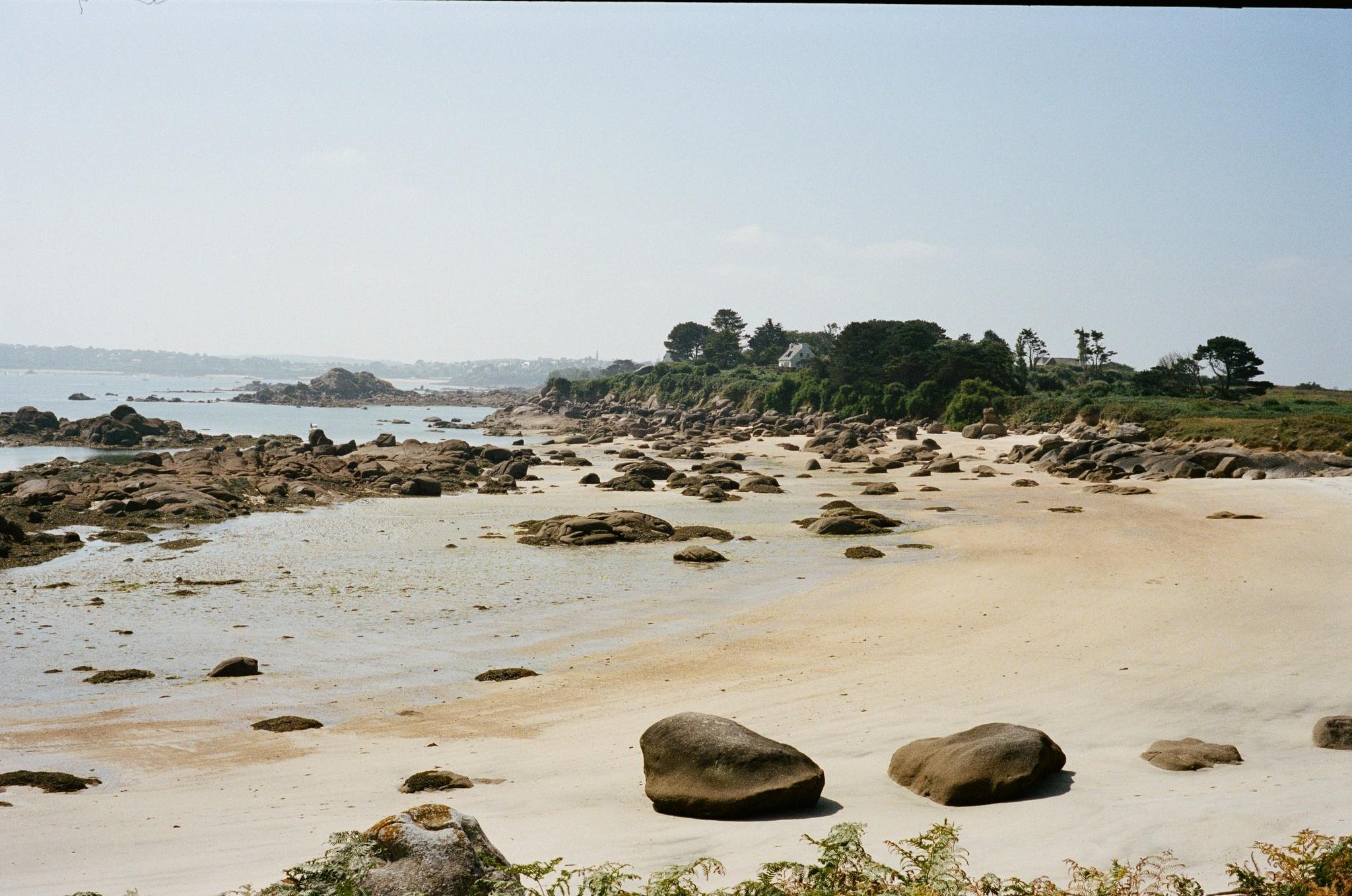 Rocky sandy beach at low tide with scattered boulders, shallow tidal pools, and a tree-lined point with a small house in the distance under a pale hazy sky.