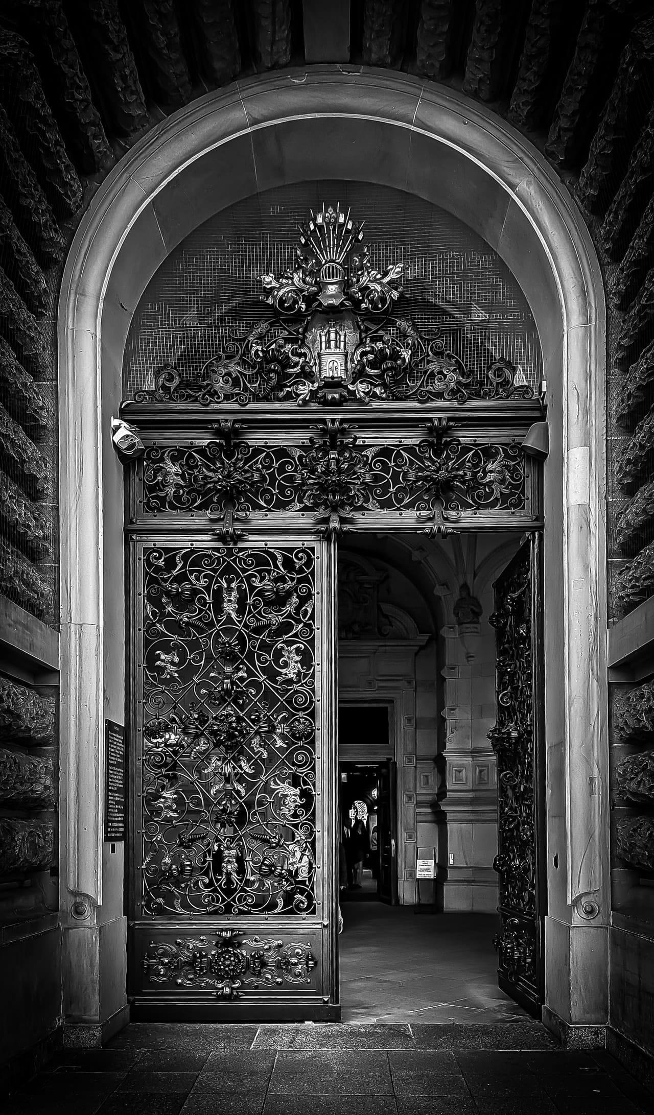Black-and-white view of a tall arched stone doorway with ornate wrought-iron double gates; one gate is open, revealing a dim interior corridor and a person standing in the distance, with a decorative crest above the arch.