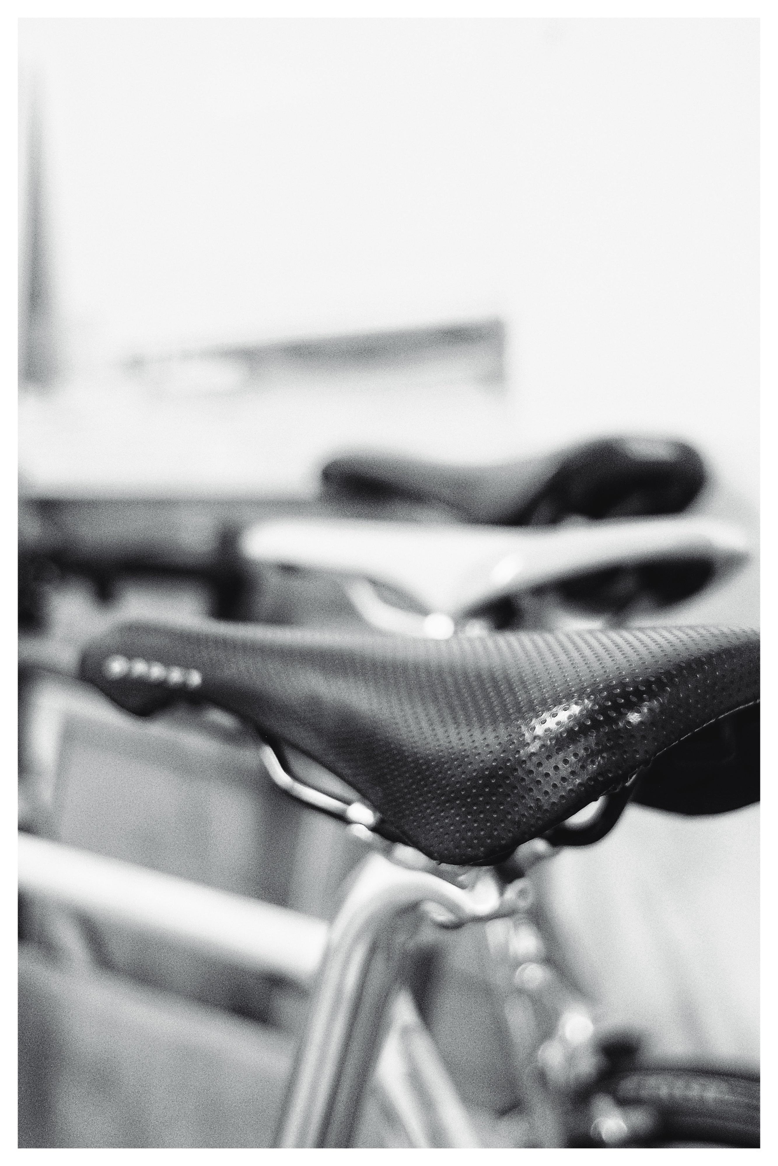 Close-up of a bicycle saddle with textured black cover and metal rails, with a second saddle blurred in the background in a black-and-white photo.