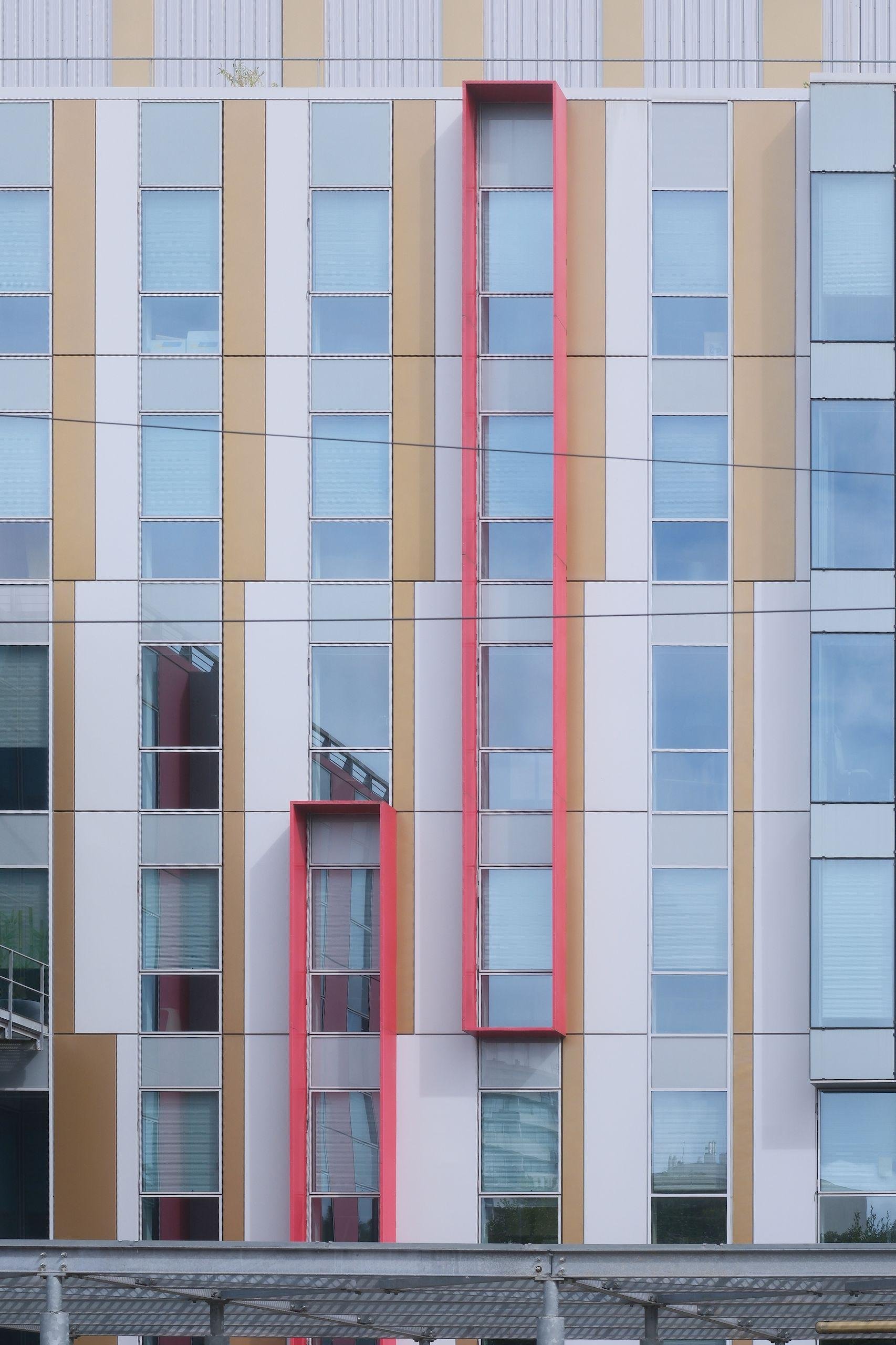 A modern building with many windows, each column being separated by light brown and white wall. Some column are framed with a red stuff (maybe to protect from the sun?) but not regularly. We can 2 windows of a column being red framed, and at the one of the top of the frame, the column on its right start to frame windows